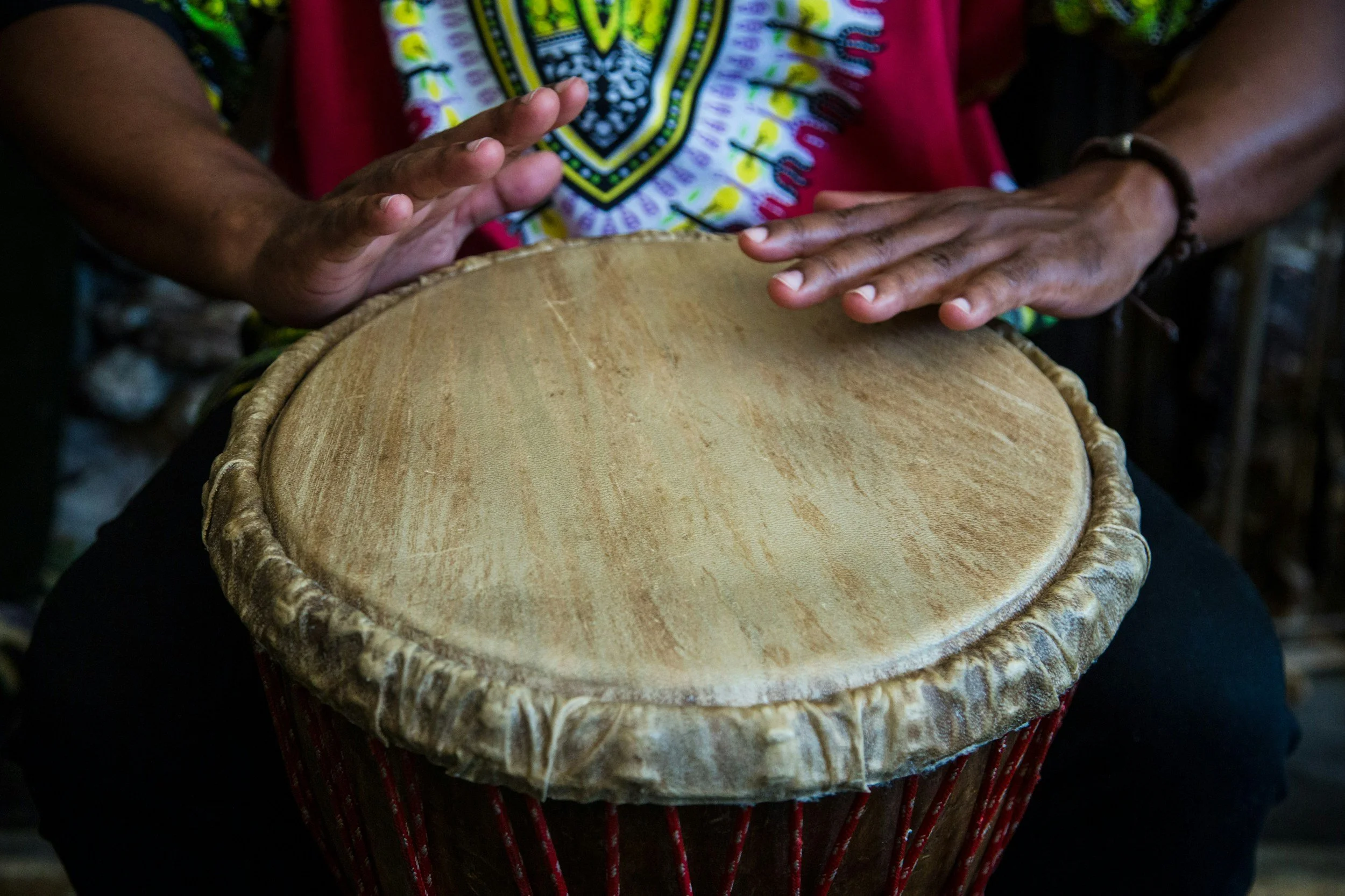 Person playing a djembe drum, focusing on hands and drum surface, wearing colorful clothing.
