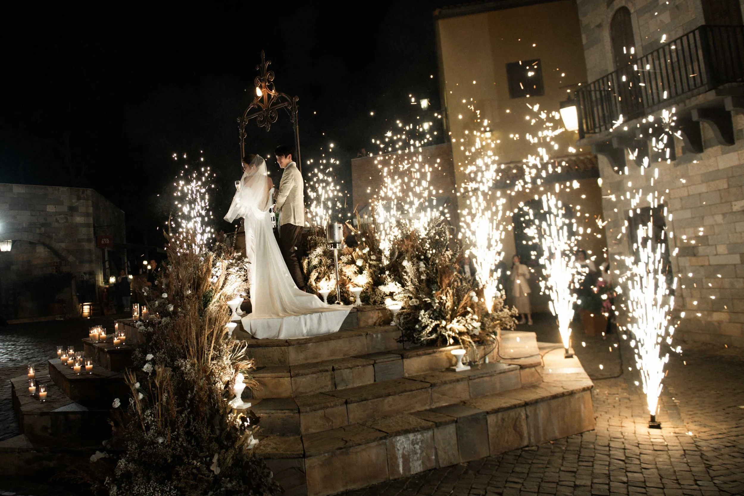 Bride and groom pouring champagne surrounded by lights and sparklers.