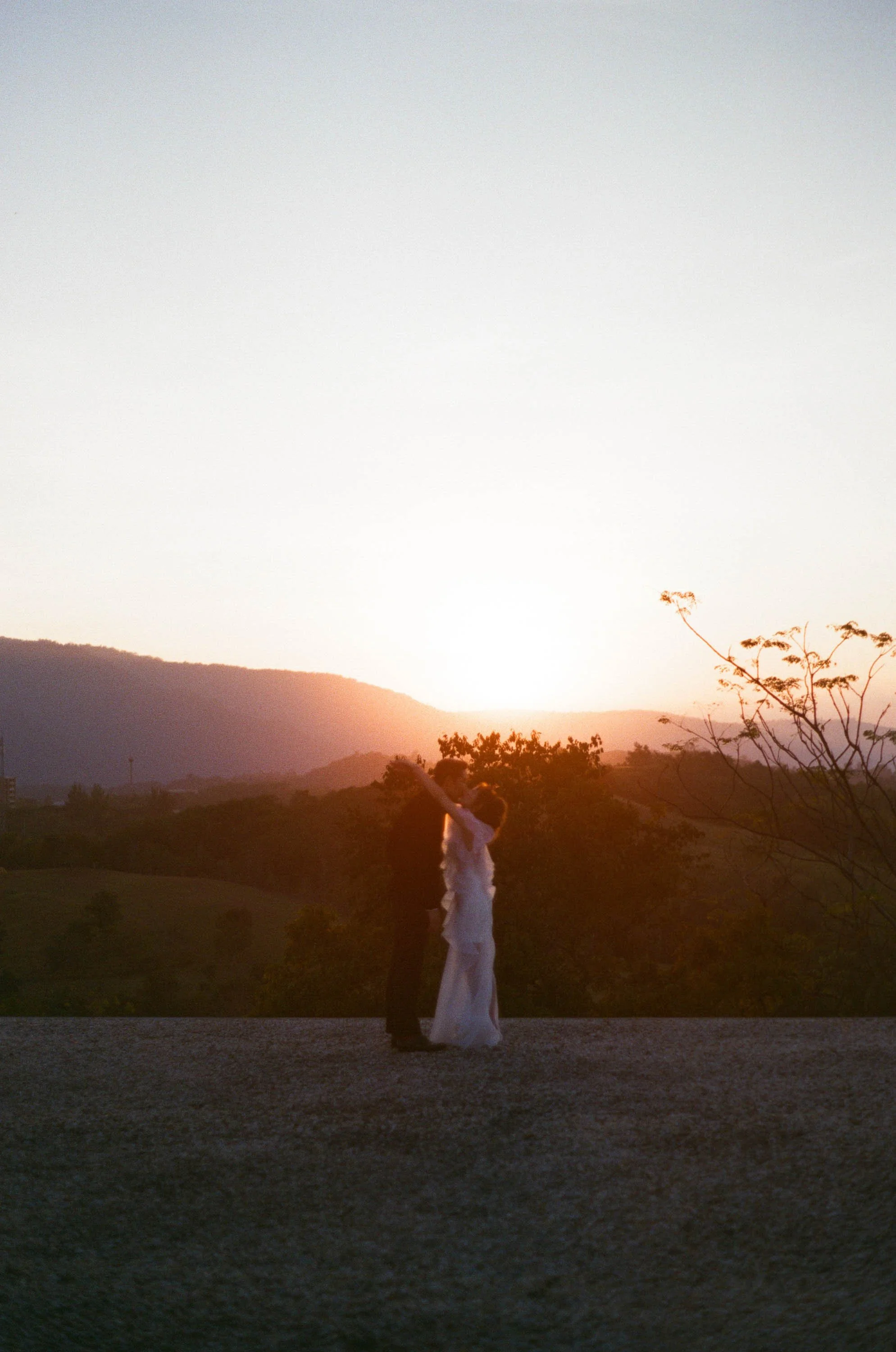 Bride and groom kissing at sunset with mountains and trees in the background.