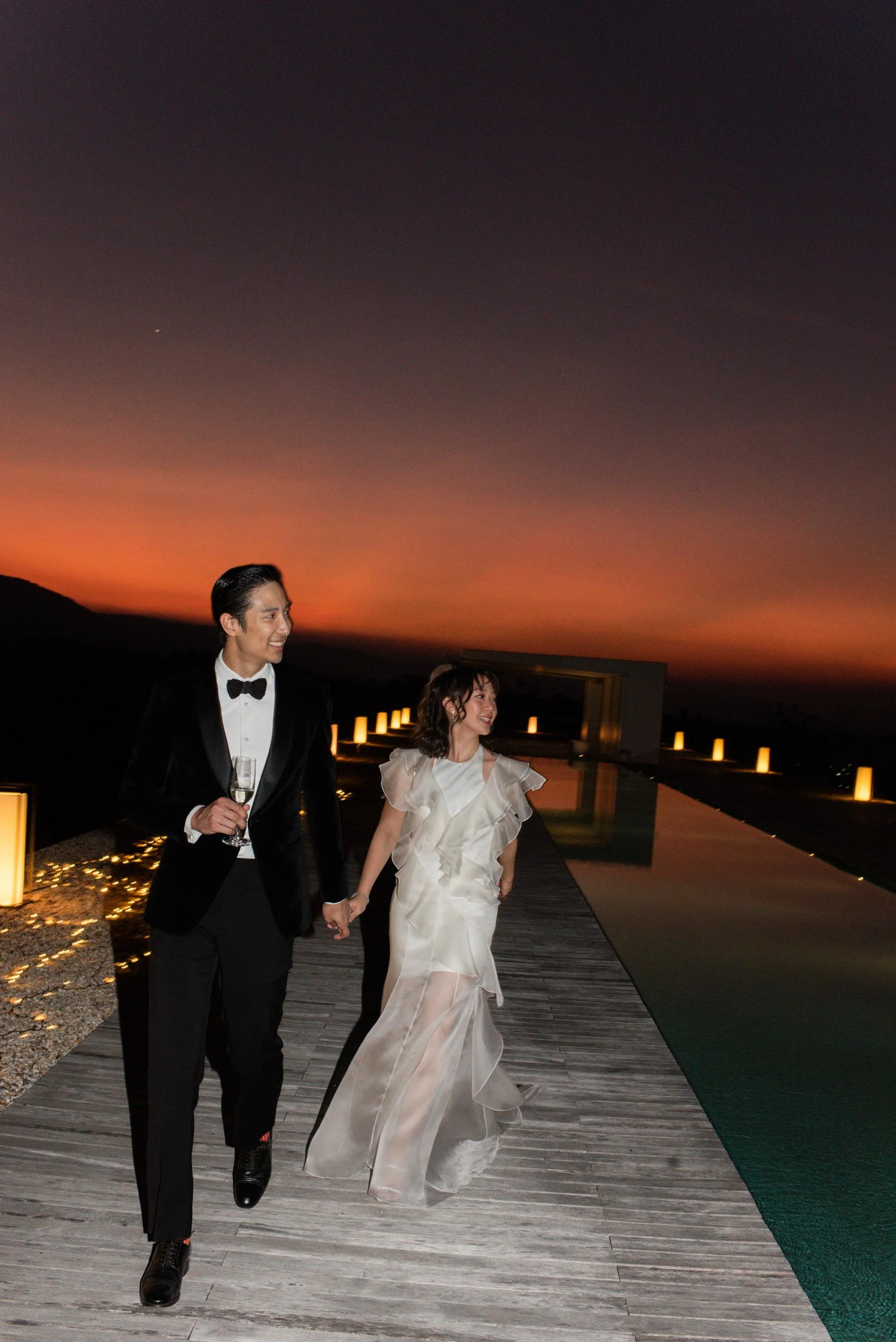 Bride and groom walking past the pool toward the dinner table at dusk.