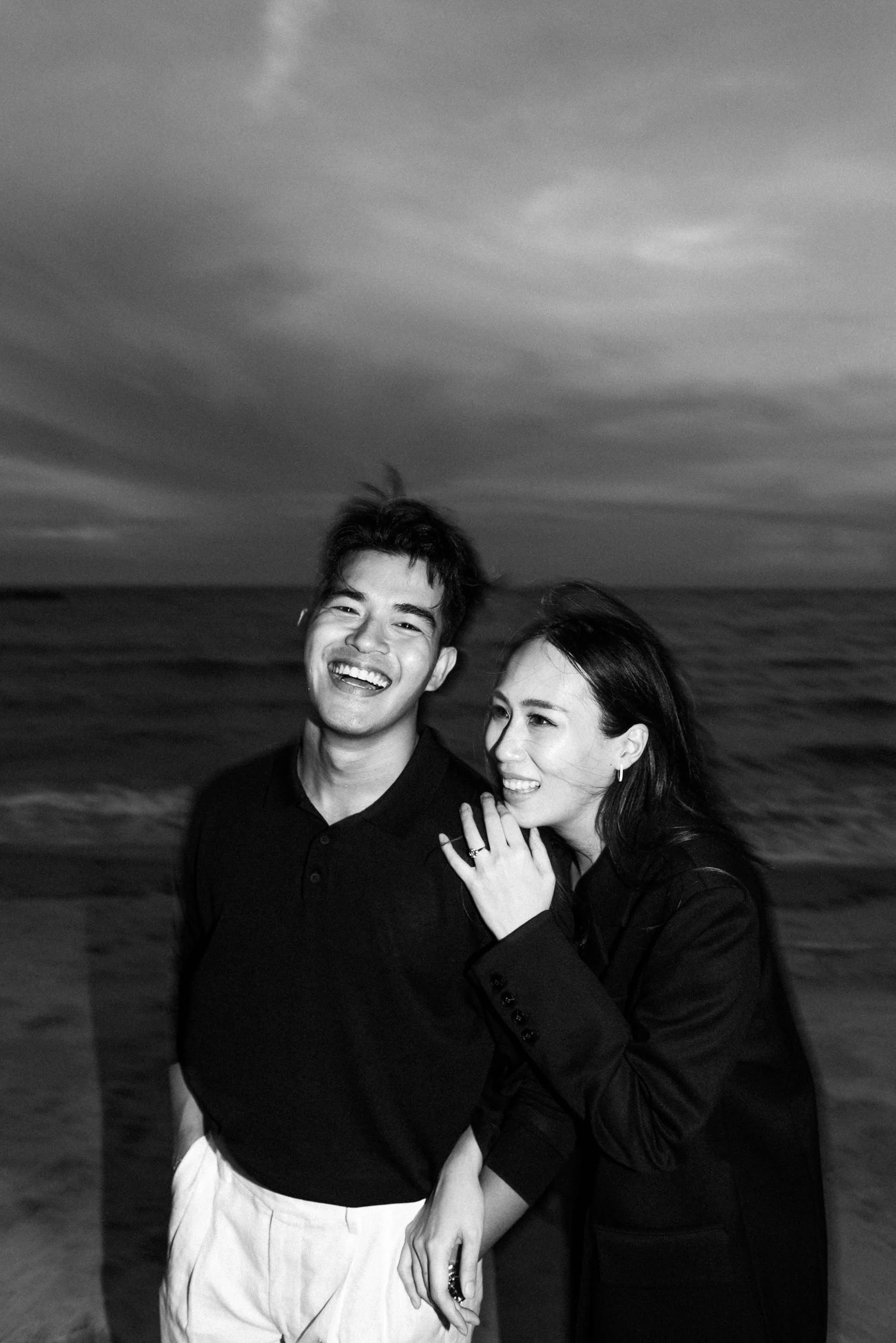 Couple in black outfits laughing and talking together by the sea.