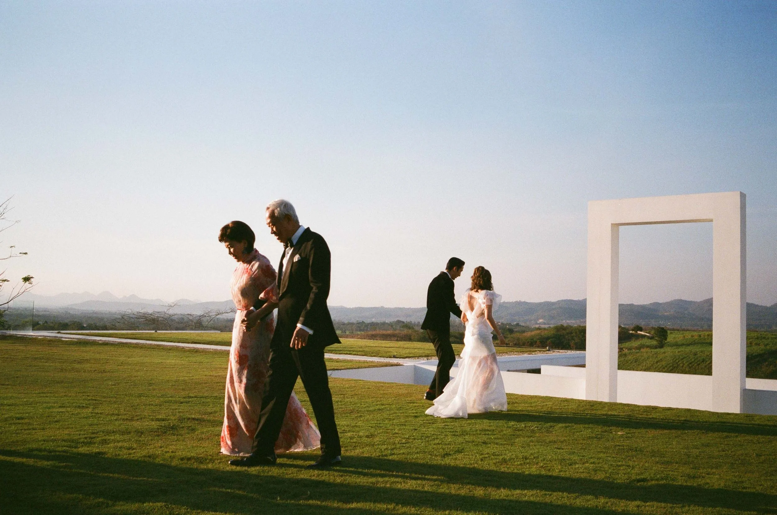 Bride and groom walking across the lawn as their parents pass by.