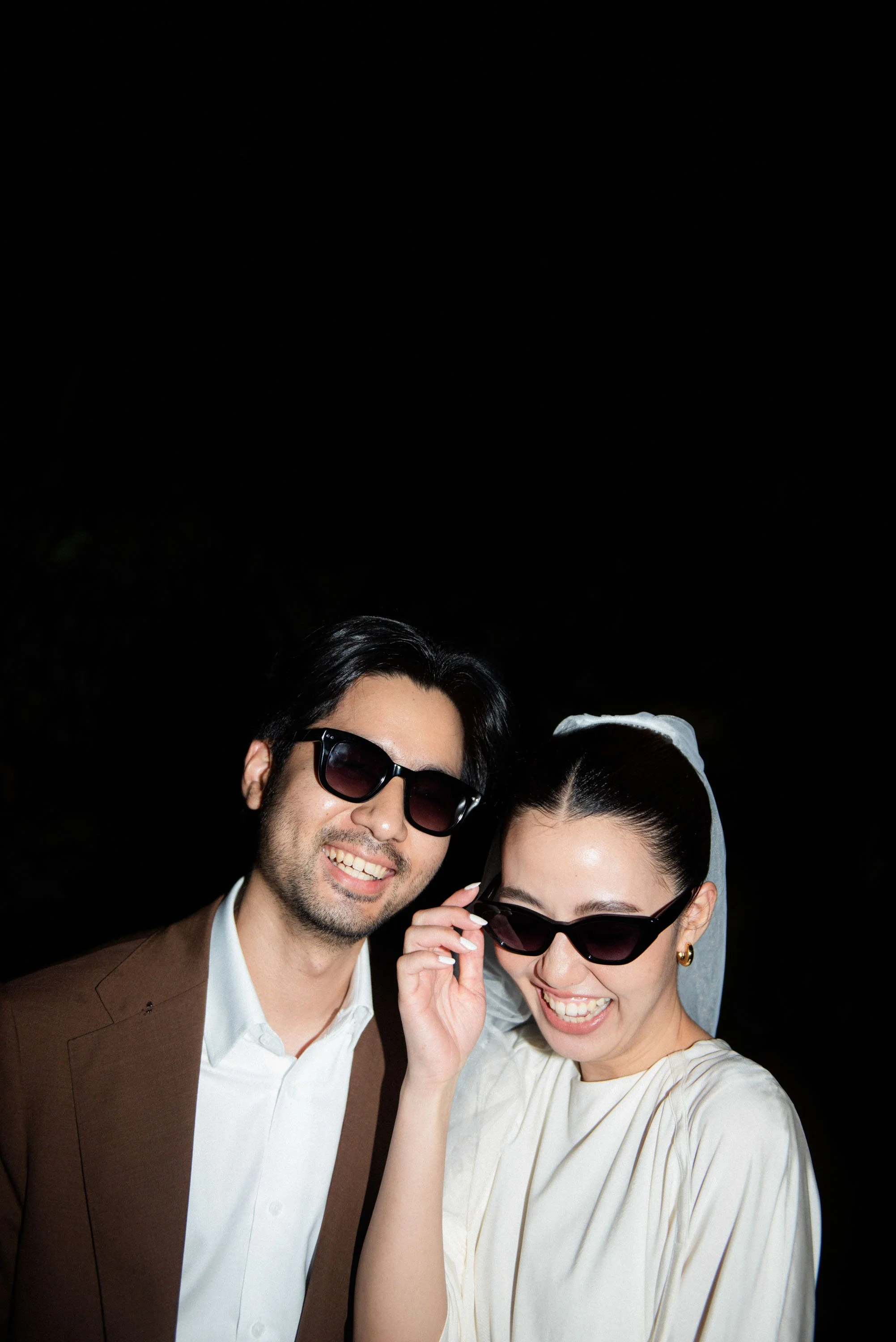 Couple standing close together wearing sunglasses and laughing during a playful portrait.