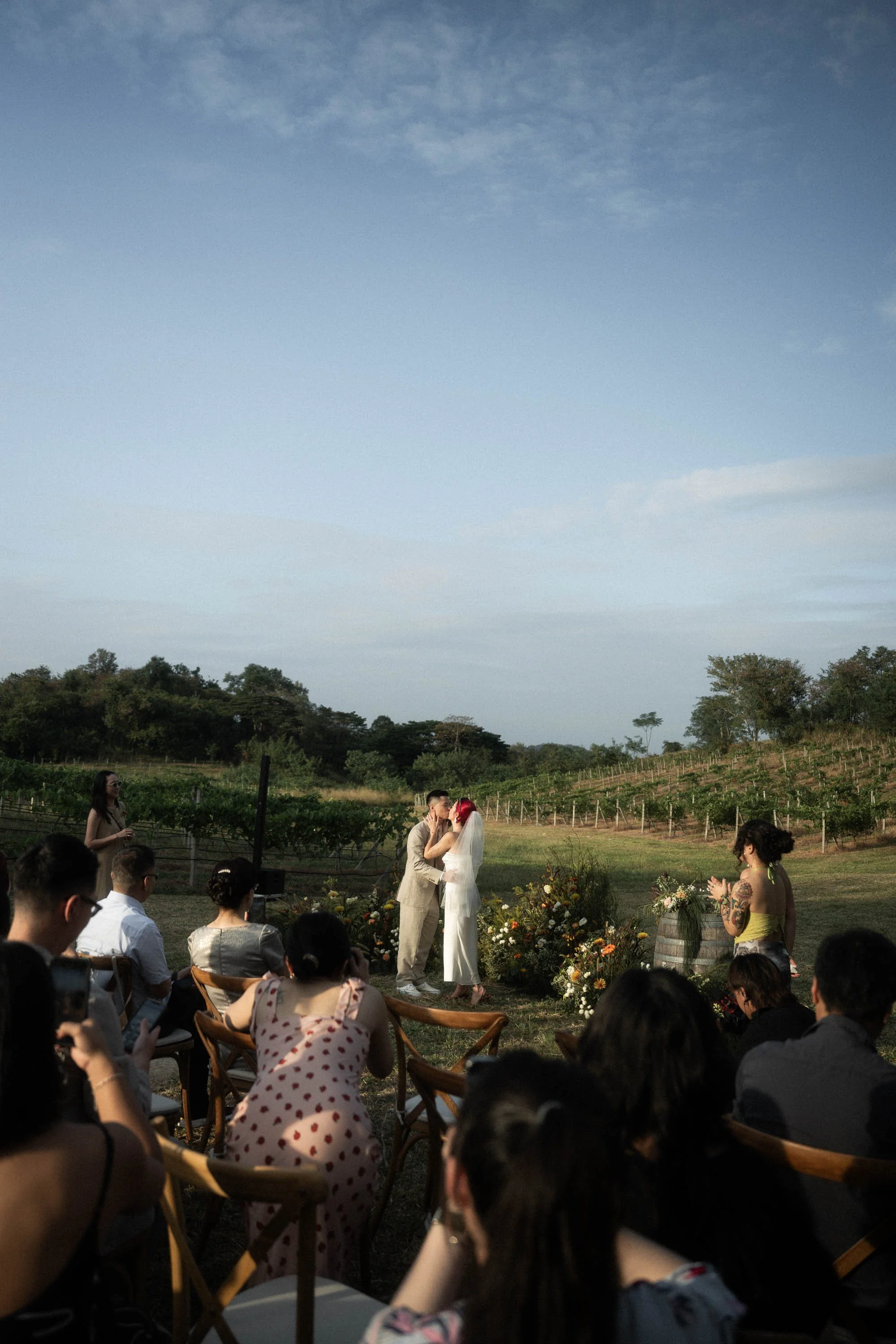 Bride and groom sharing a kiss after the wedding vows with guests and the vineyard in the background.