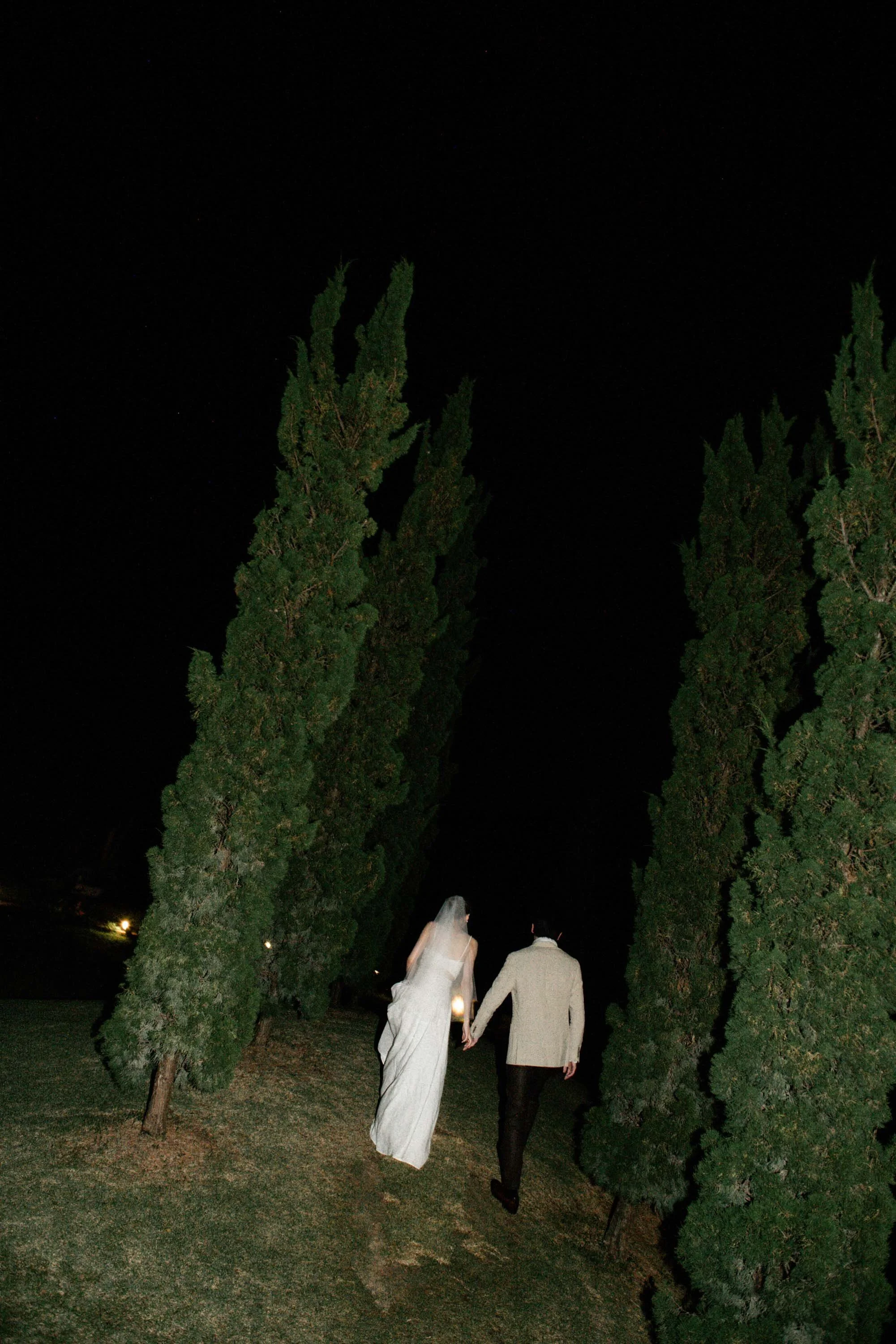 Bride and groom walking together among the trees at night.