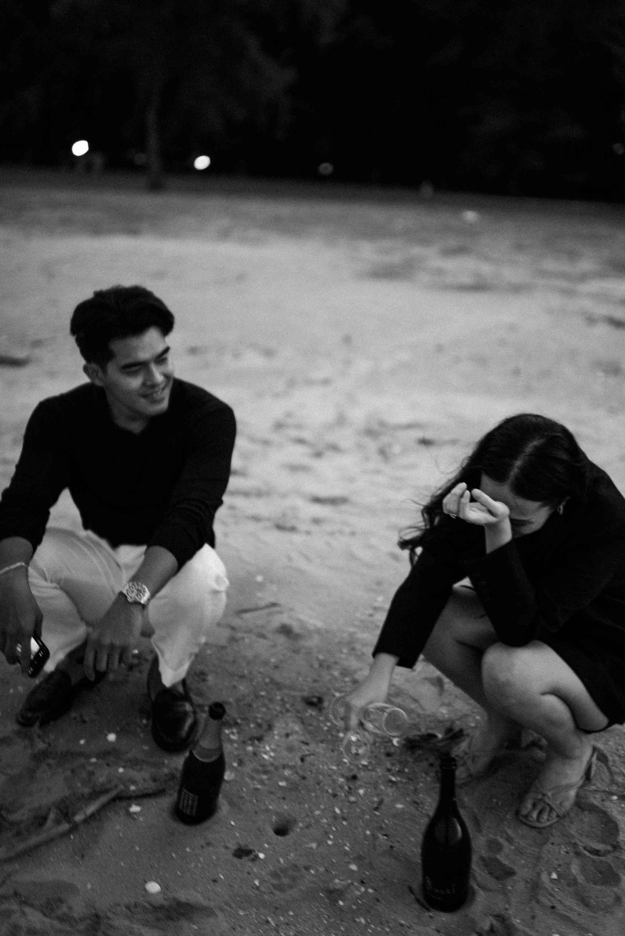 Couple crouching on the beach while opening a bottle of champagne.