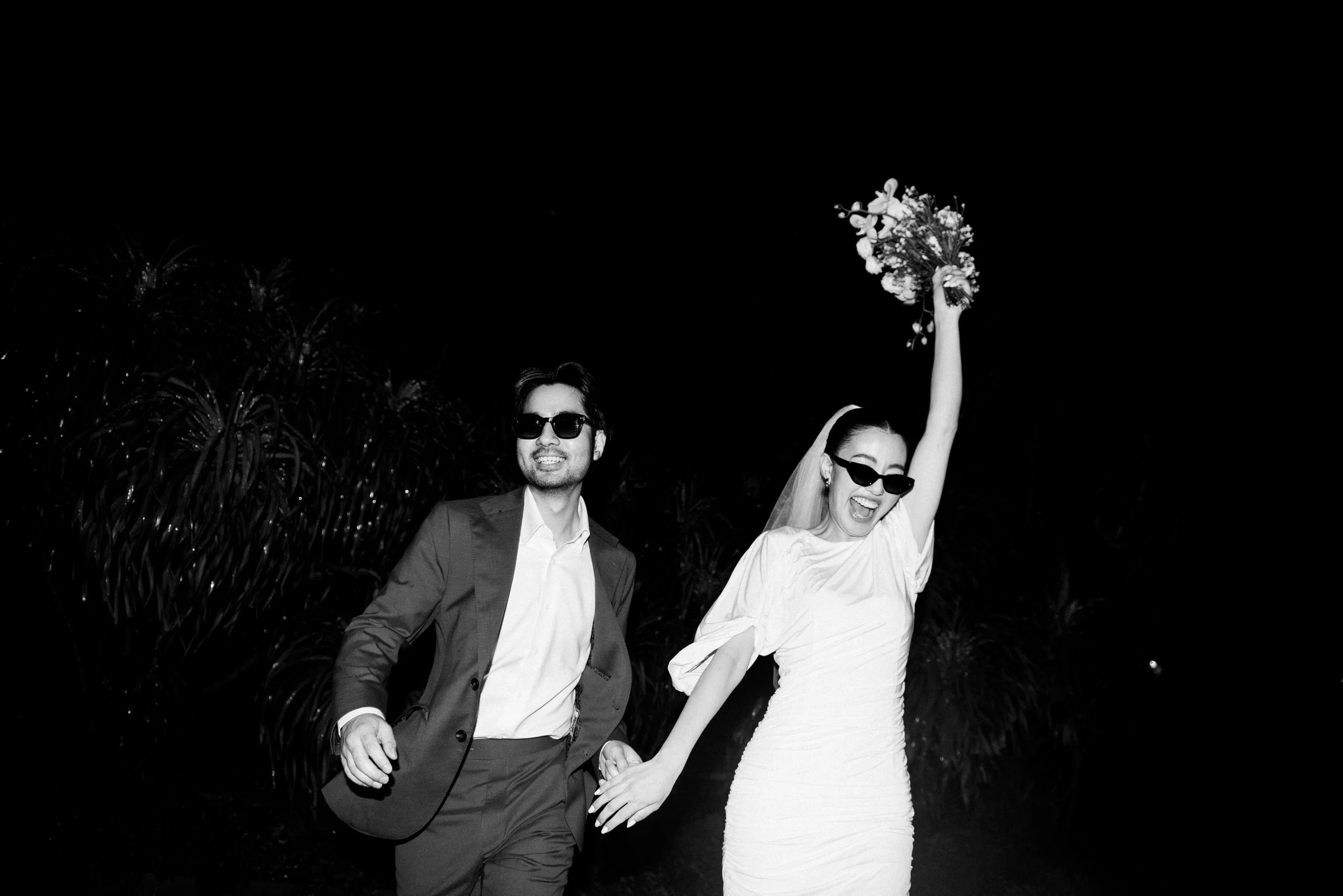 Couple running together across the garden as the woman raises her bouquet in the air.,