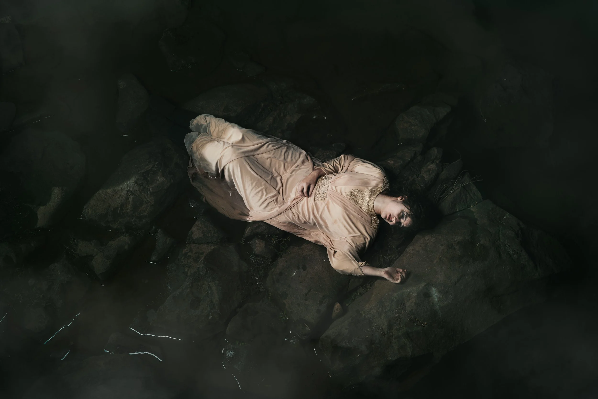 Fine art photograph of a woman in a cream dress lying on river rocks in shallow water under soft cinematic lighting.