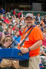 A man with a beard and glasses wearing an orange shirt and a baseball cap, standing among a crowd at an outdoor event, holding a blue box and smiling at the camera.