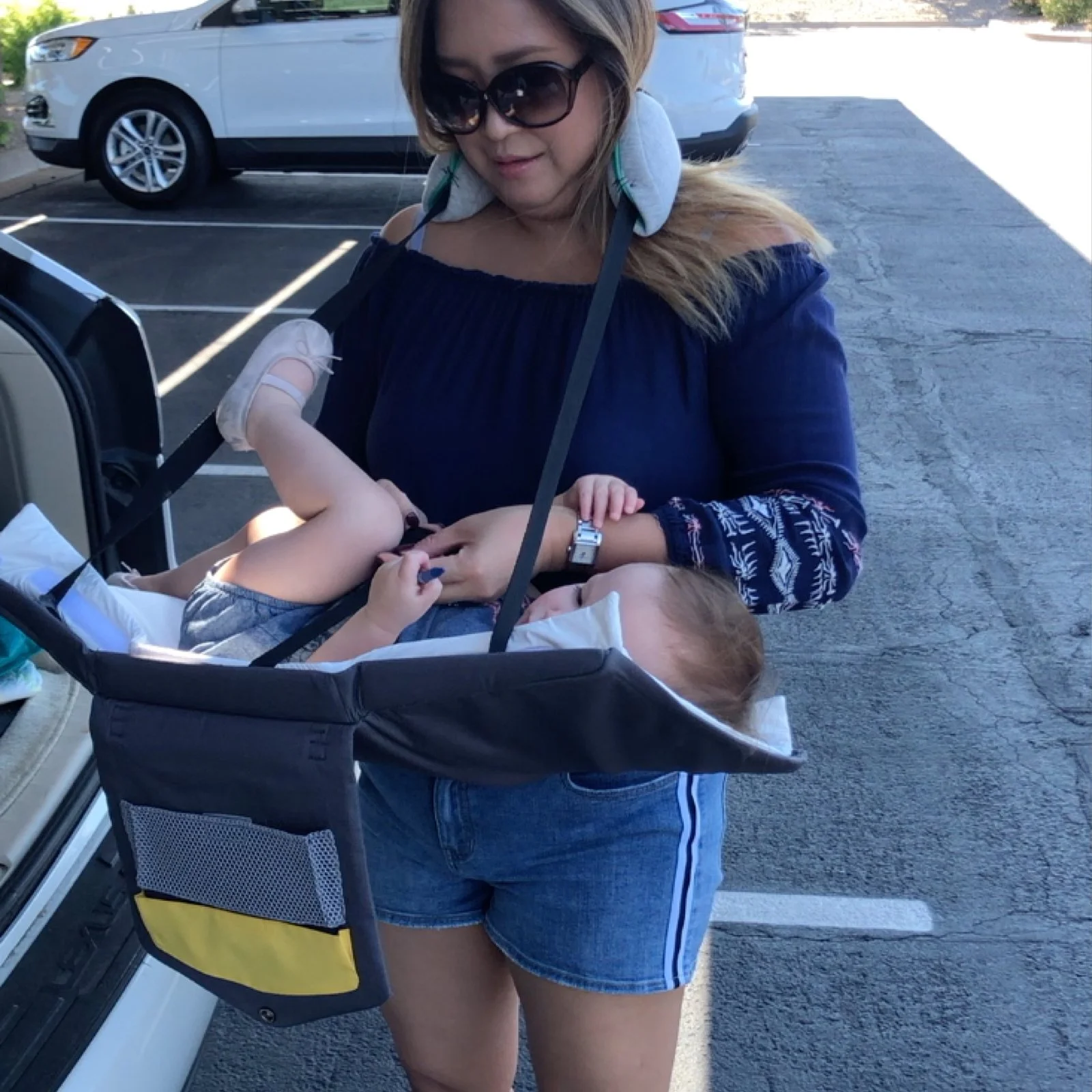 A woman wearing sunglasses and a black off-the-shoulder top is standing outside a parking lot, looking down at a young child lying in a wearable changing station. The woman is holding the child's hand.