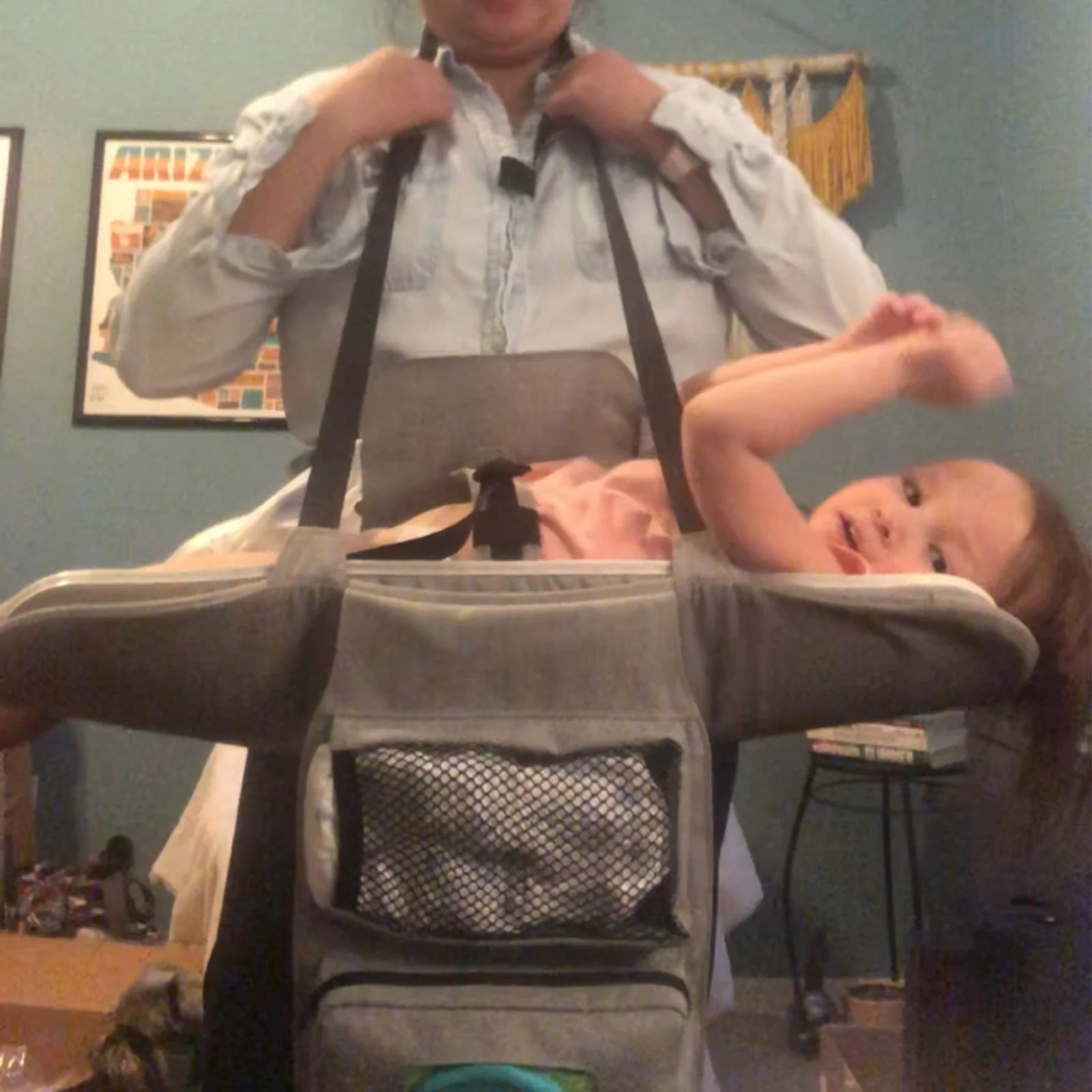 A person standing behind a dog in a stroller, with a young girl lying inside the stroller, indoors with a blue wall, framed art, and furniture in the background.