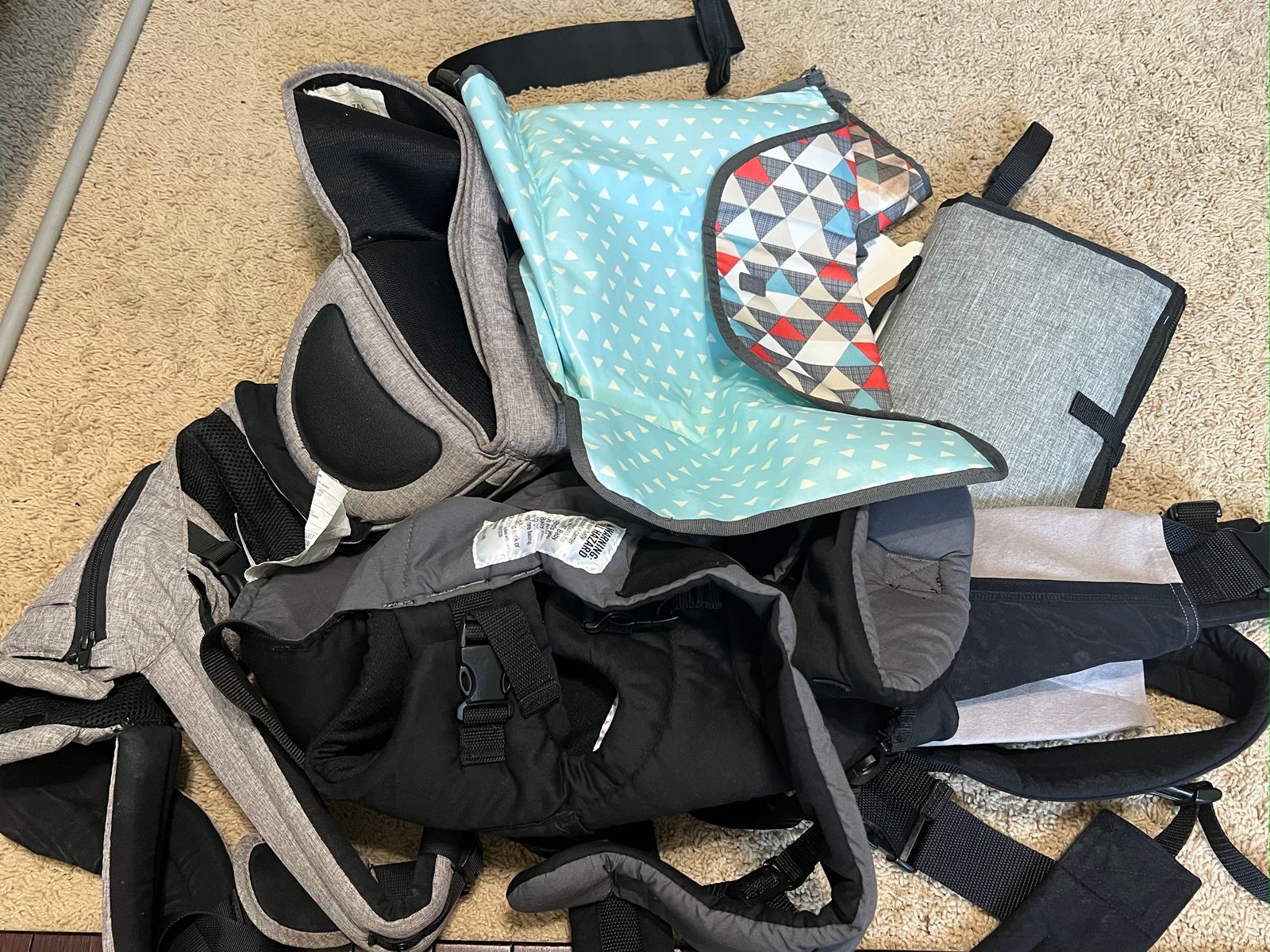 A collection of used backpacks and a diaper bag spread out on a beige carpeted floor.