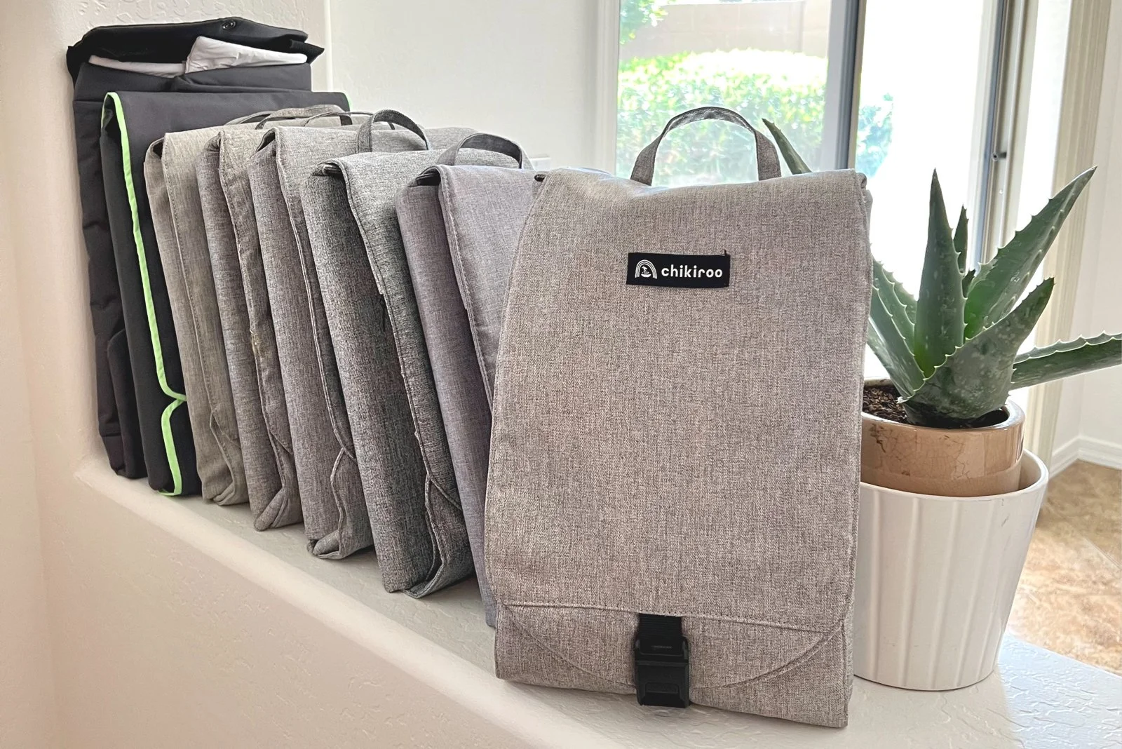 A row of gray and black fabric bags with handles, labeled 'chikiroo,' placed on a white ledge near a window, with a large potted succulent plant nearby.