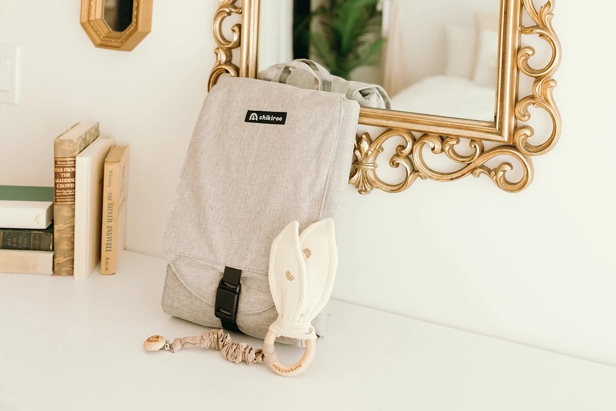 A beige fabric backpack hanging on a decorative gold-framed mirror, with a wooden bunny-shaped teething toy attached, on a white surface next to a stack of books and reflected in the mirror.