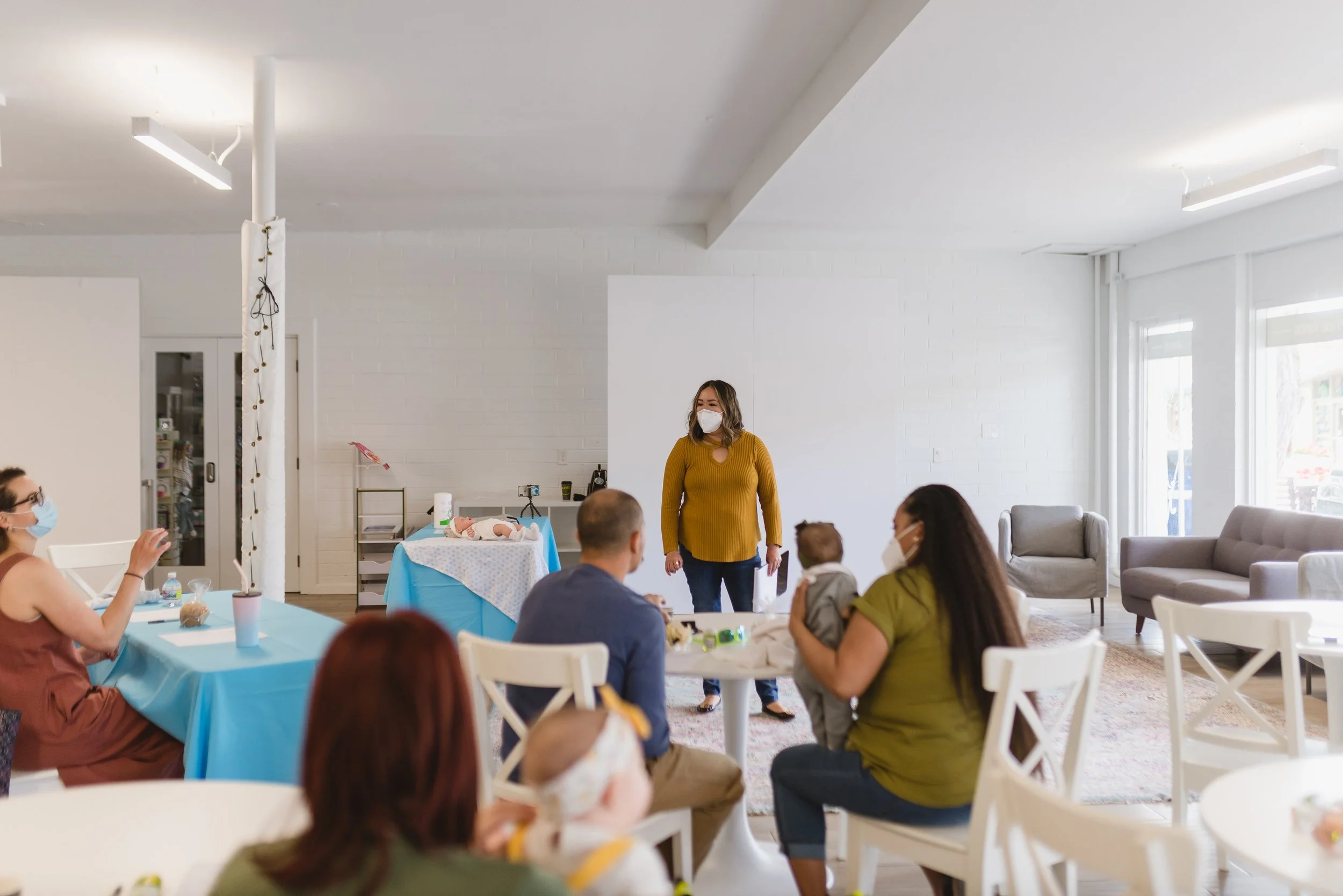A group of people, some wearing masks, attending a baby shower in a bright, modern room with white walls and large windows. A woman in a mustard yellow sweater stands near a table with a doll for a baby doll diaper changing activity. There are tables with blue tablecloths, snacks, and drinks, and a decorative pole with string lights.