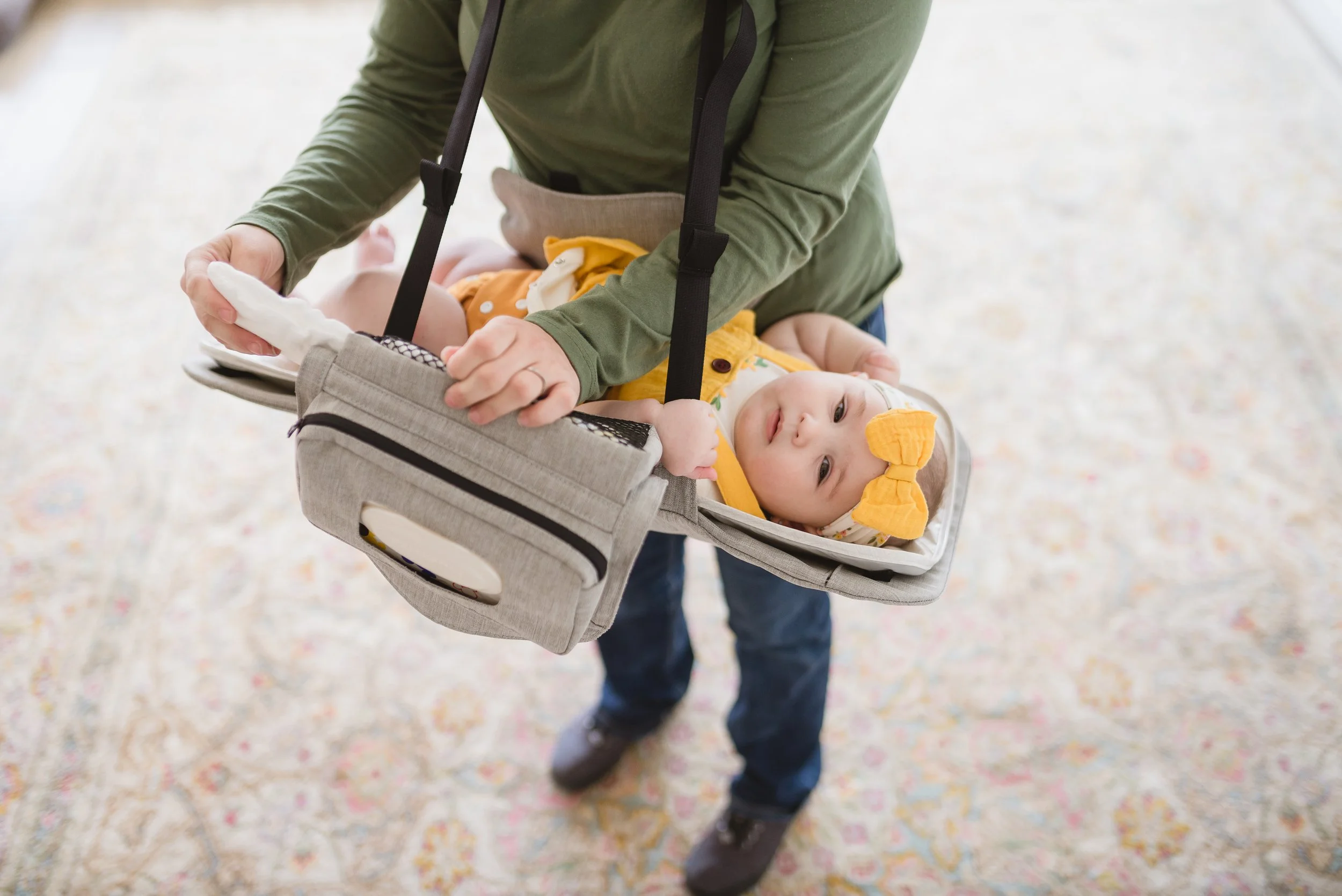 Person carrying a baby in a car seat at an indoor location with a colorful carpet.