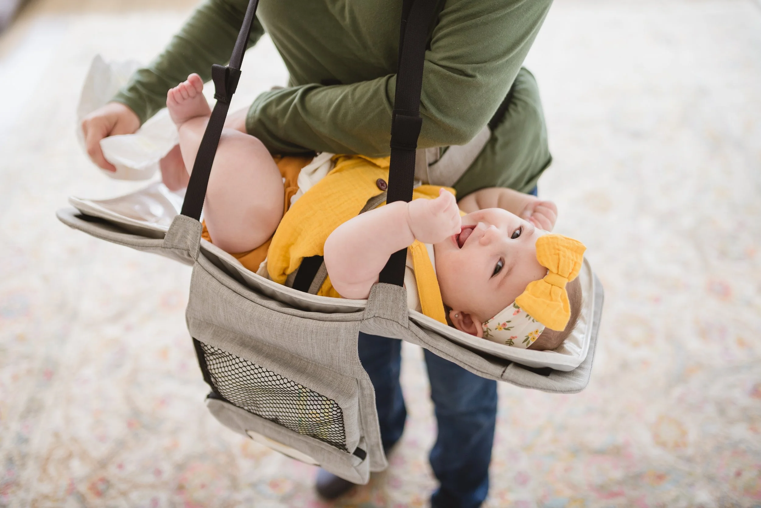 Child lying on a swing in a stroller, smiling and looking up at an adult who is pushing the stroller. The child is wearing a yellow hat with bear ears, a yellow shirt, and floral-patterned bottoms.