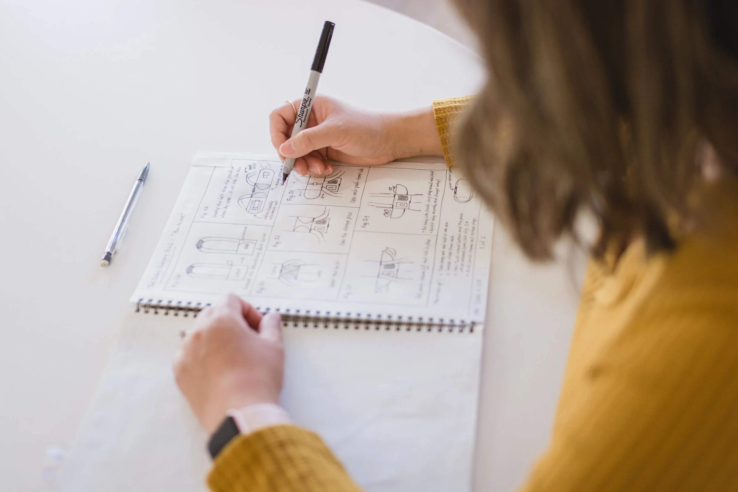 A person writing on a notebook with hand-drawn diagrams and notes, with a pen placed on the white table and another person's hand holding the notebook.