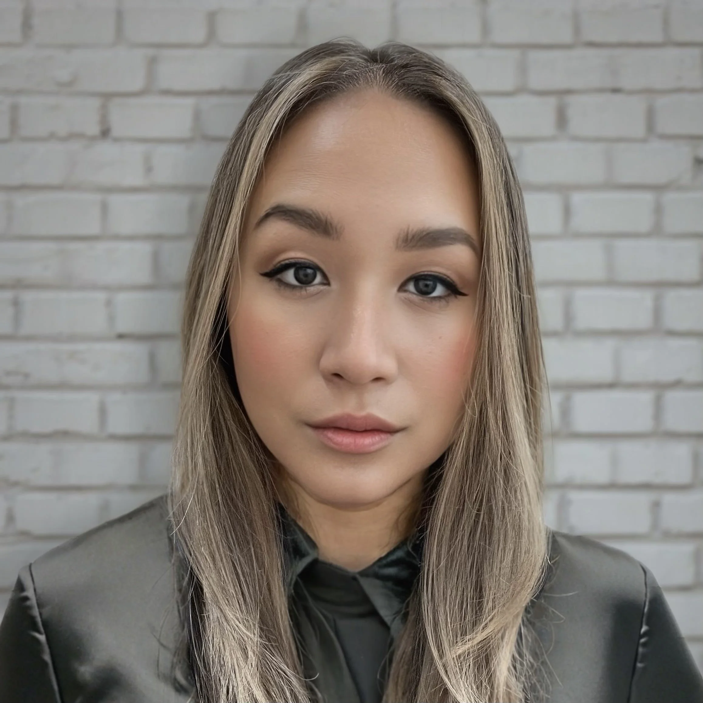 Close-up of a woman with blonde hair and makeup, standing against a gray brick wall.