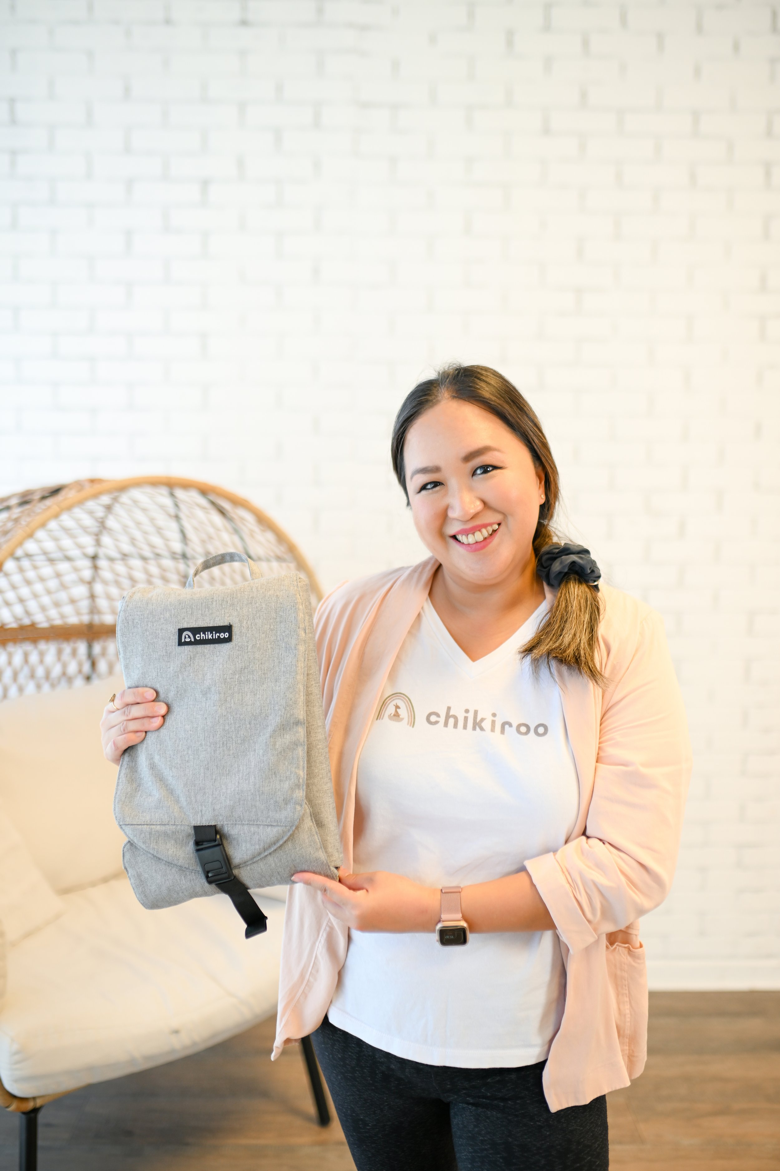 A woman with long hair tied in a side braid smiling and holding a gray backpack with a brand label, standing in front of a white brick wall indoors.