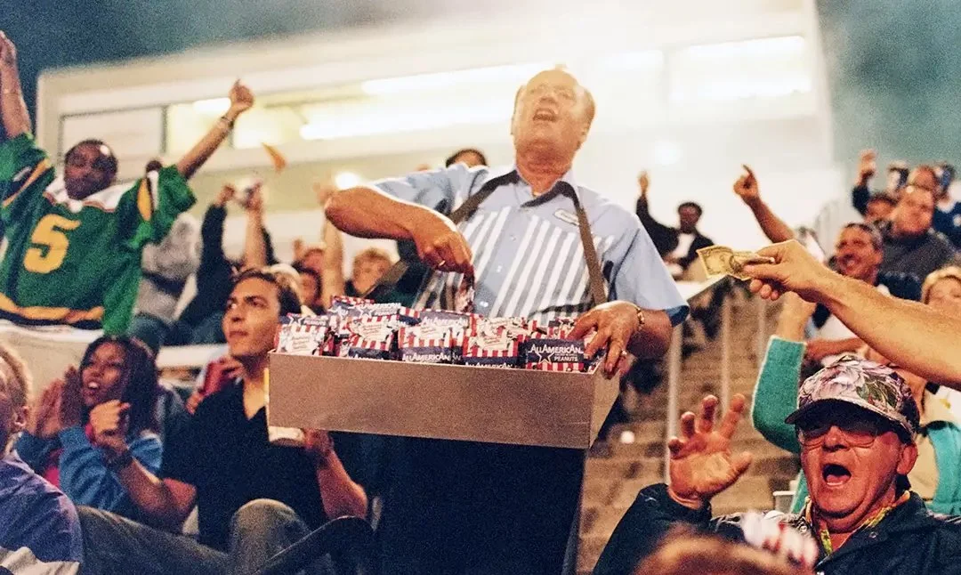 A crowd at a sports event, with people cheering and one man holding a tray of snacks or treats, while another person hands him cash for payment.