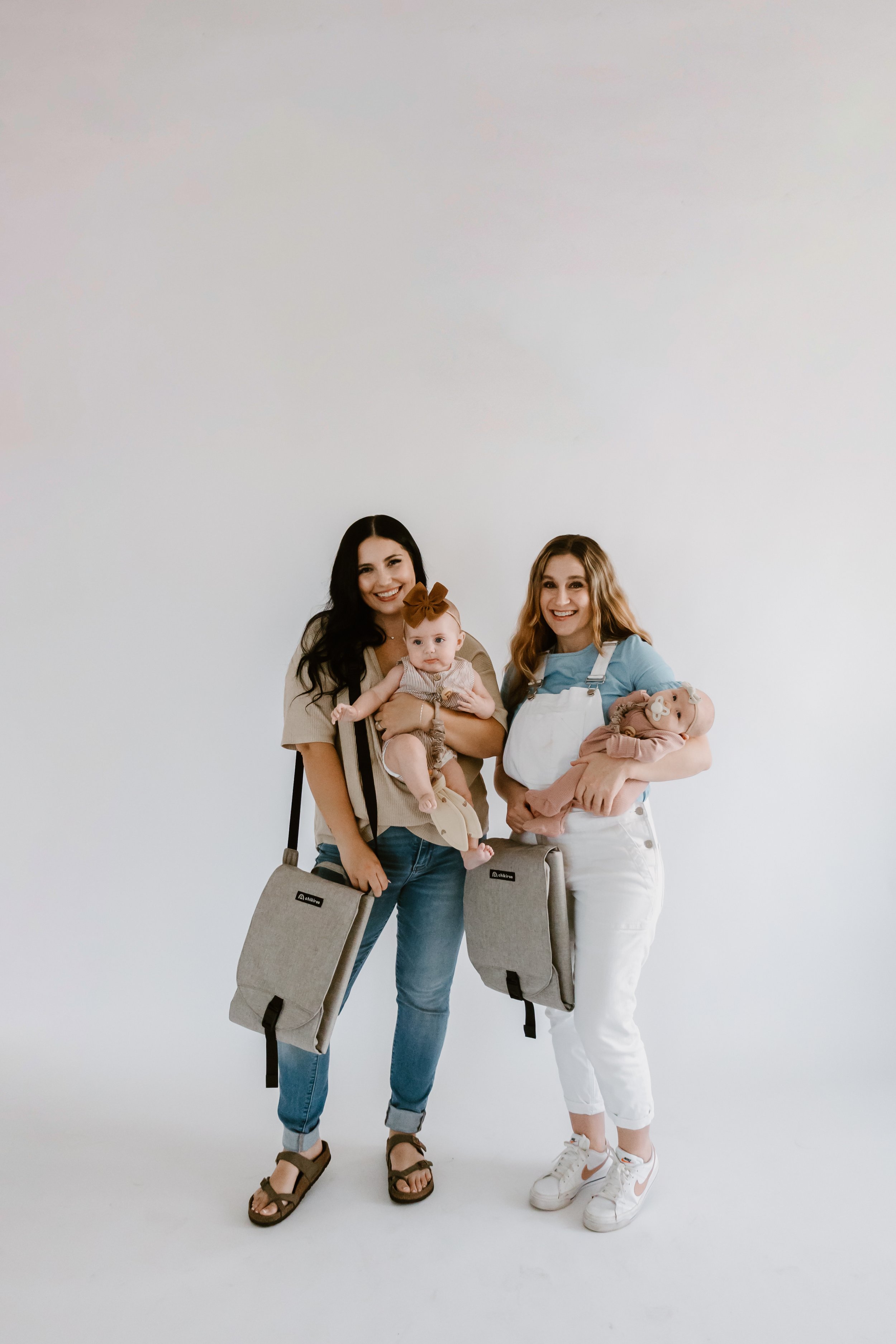 Two women holding babies, standing against a plain white background, smiling at the camera. Each woman has a portable movie or TV screen on a strap hanging from her shoulder.