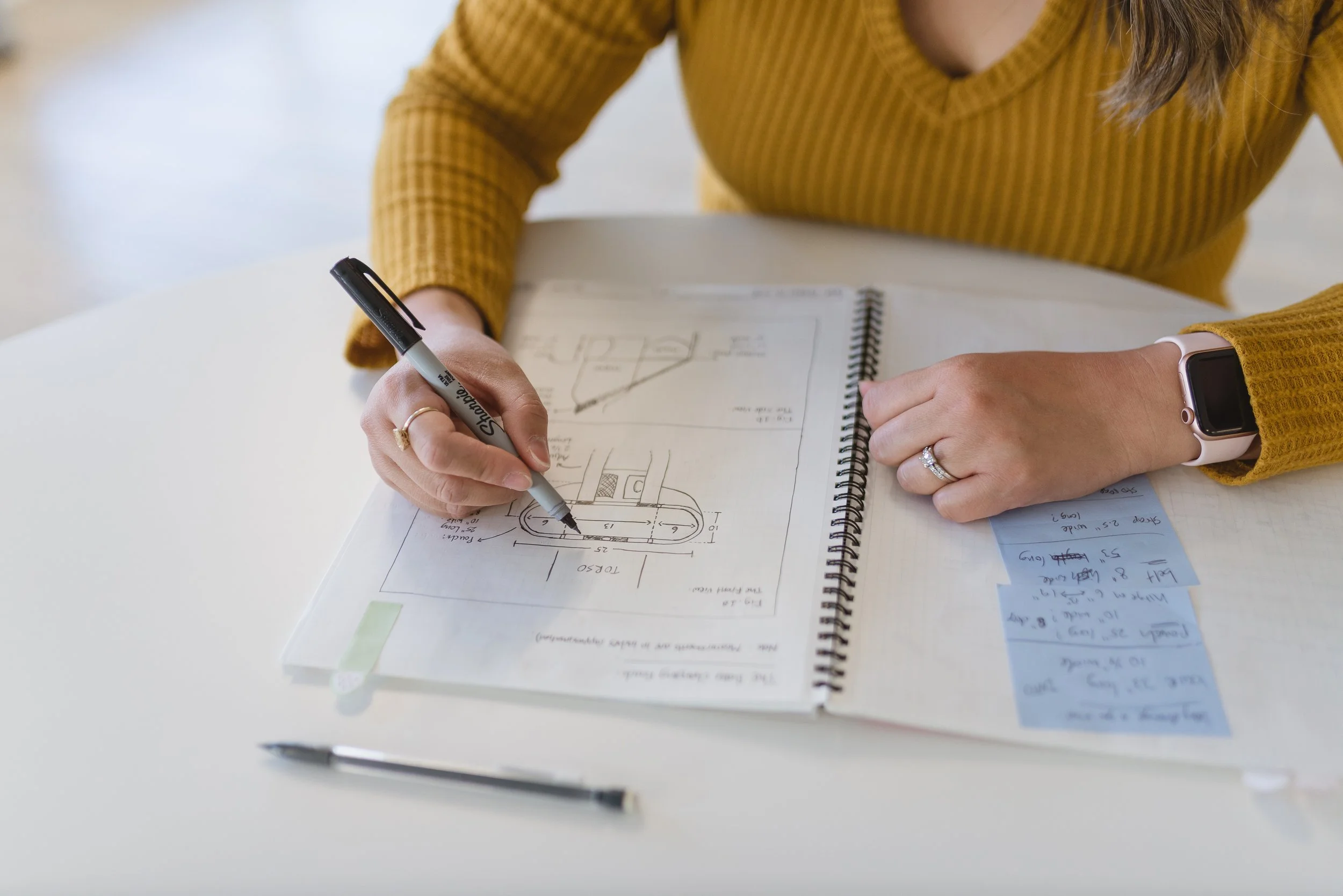 A person wearing a mustard yellow sweater is writing with a black marker on architectural plans in a spiral notebook, placed on a white desk.