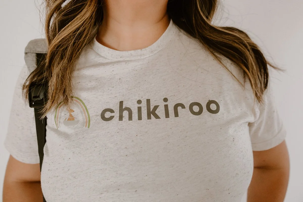 A woman wearing a light gray t-shirt with the word 'chikiroo' printed on it and a small rainbow logo, with a black strap of a backpack visible on her shoulder.