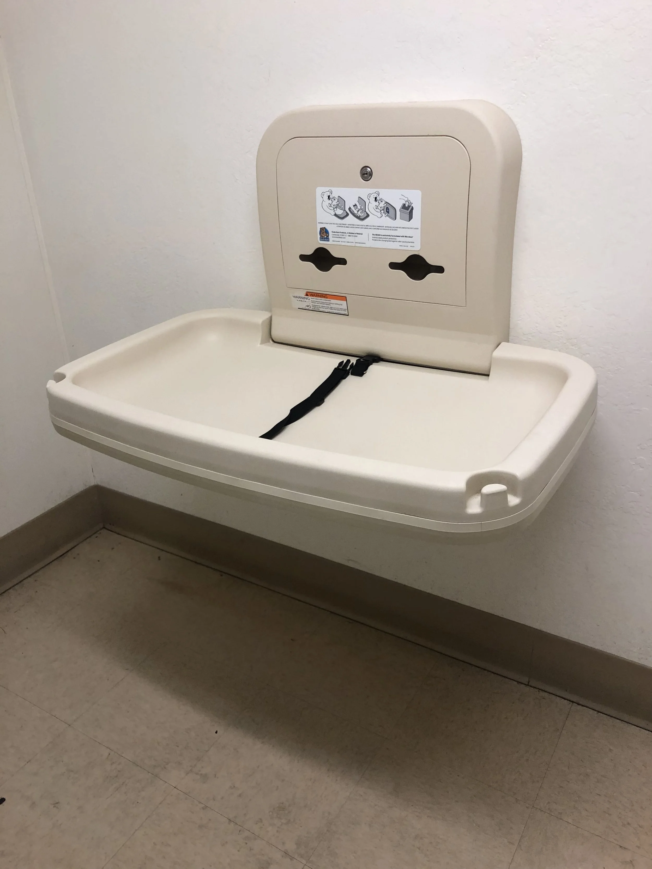 A beige plastic changing station mounted on a white wall in a public restroom.