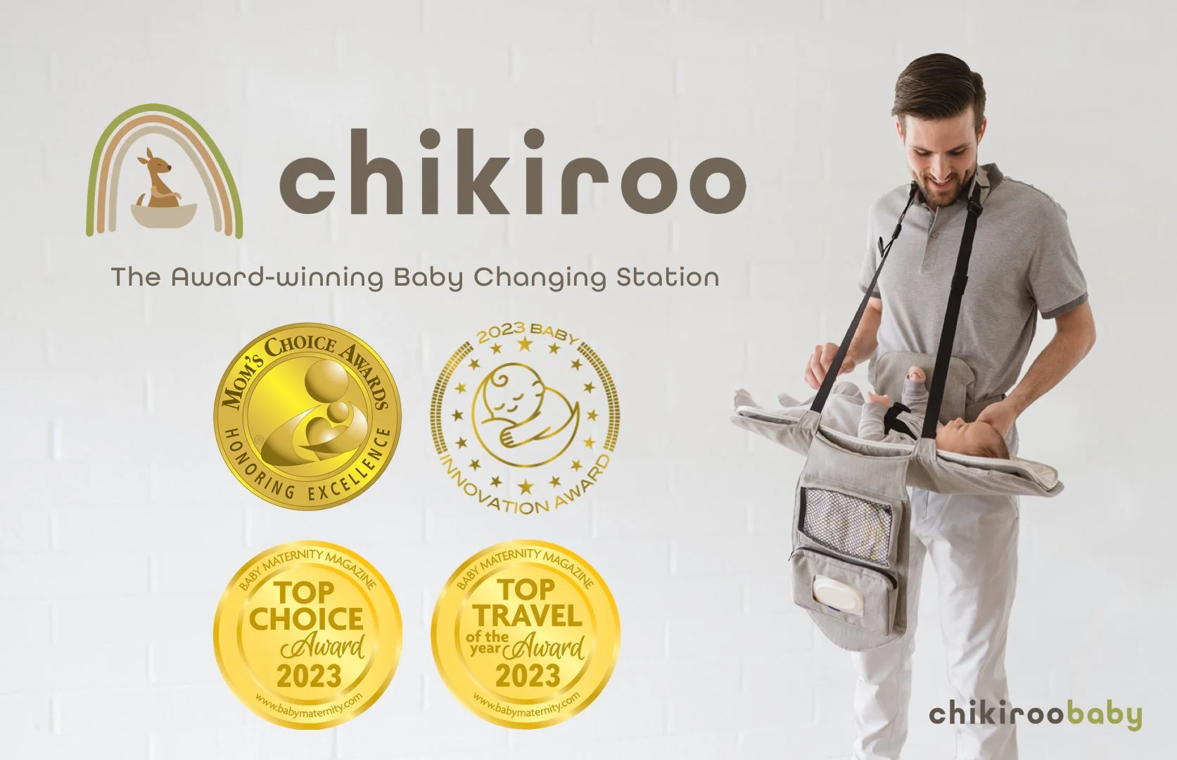 A man with a beard smiling and looking down at a baby in a baby carrying bag attached to his chest. The baby is lying in the bag, and the man is holding the bag with both hands. The background features a white wall with various award medals and a logo that reads "chikiroo" and "The Award-winning Baby Changing Station."