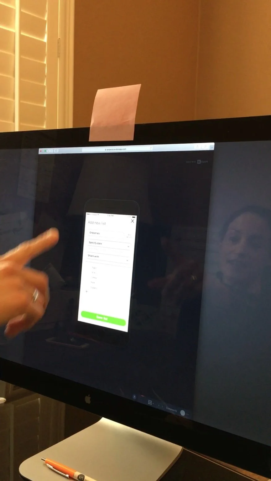 Person pointing at a smartphone screen displayed on a computer monitor, showing a digital list creation interface with options for groceries, date, and sharing. There is a pink sticky note attached to the top of the monitor, and a pen resting on the desk below.