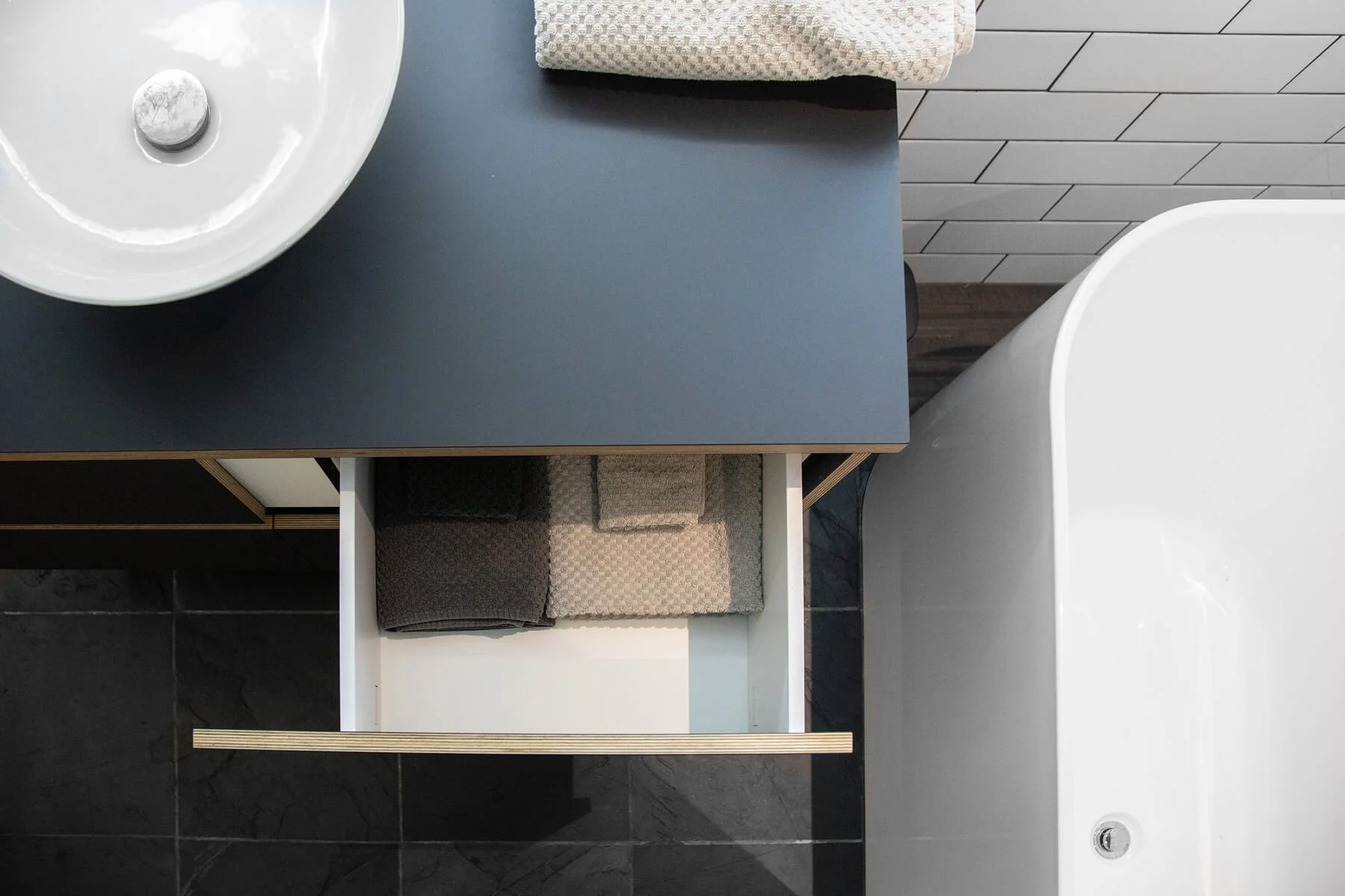 Bathroom with a white sink, a dark gray countertop, a towel, and a storage shelf with towels and washcloths. The wall is tiled with white rectangular tiles, and the floor has black marble tiles.