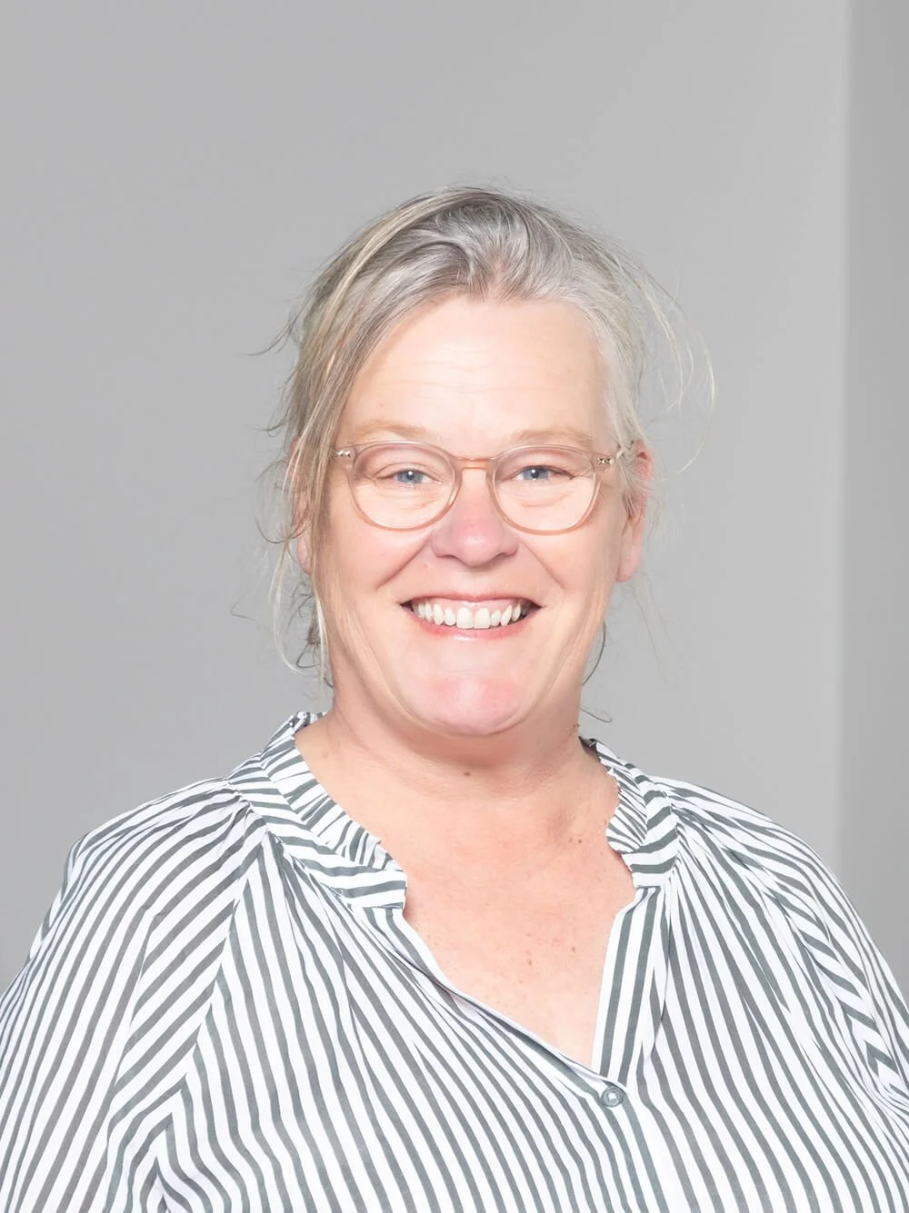 A smiling middle-aged woman with glasses and gray hair, wearing a black and white striped blouse and black pants, standing in front of a light gray wall with a potted plant in the background.