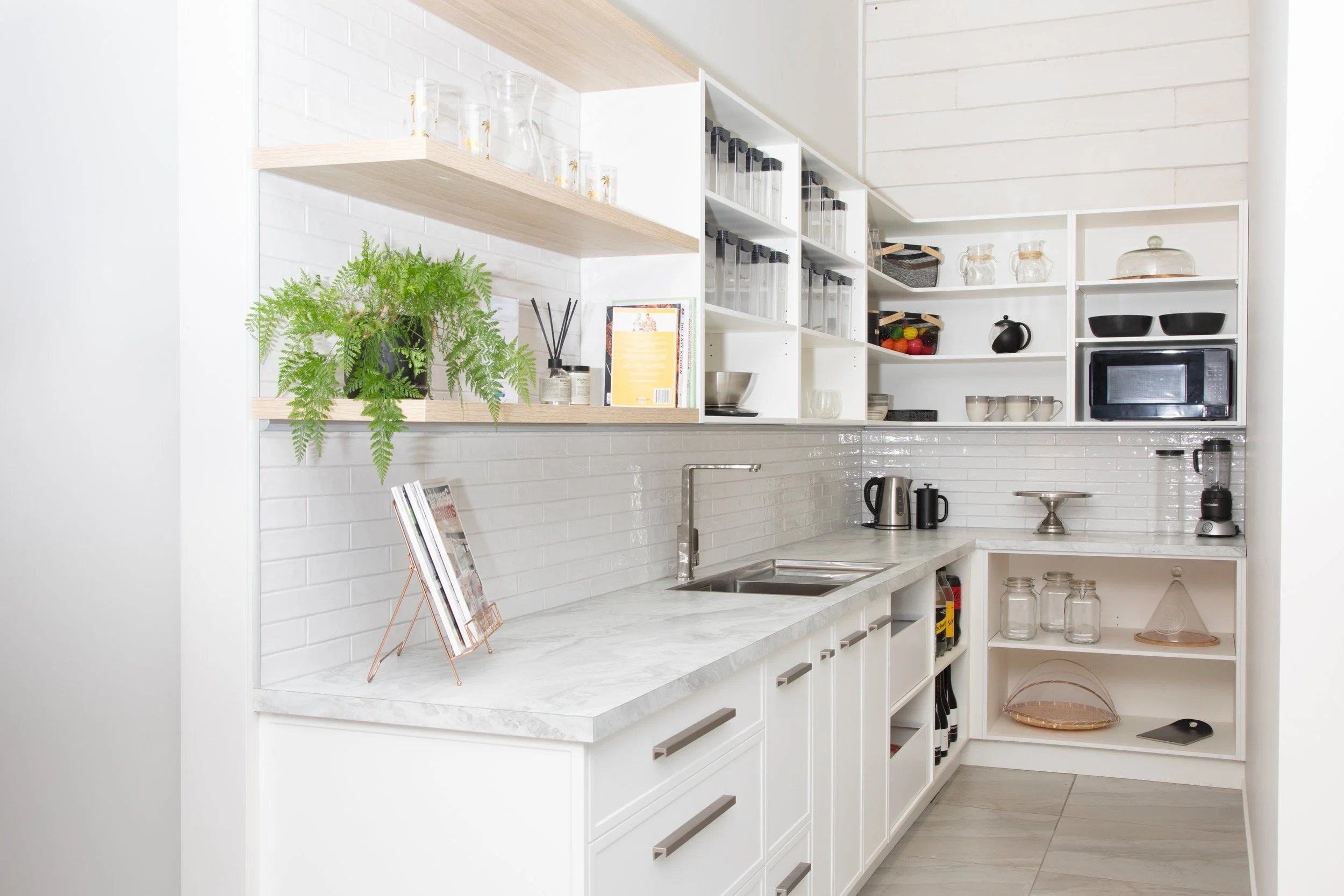 A modern kitchen with white cabinets, open shelving, and a marble countertop, decorated with a list of books, green plant, and kitchen appliances.