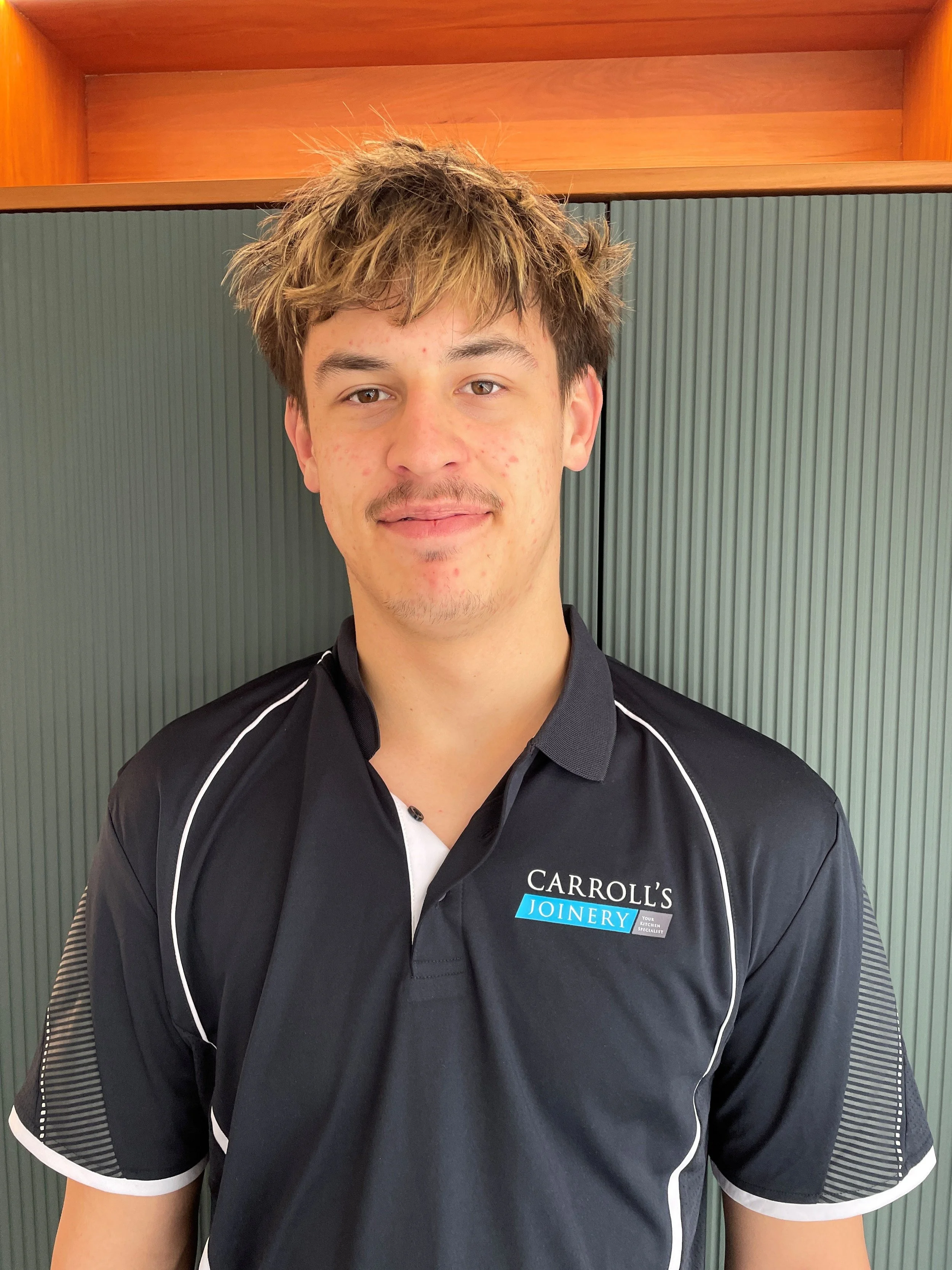 Young man with short, tousled brown hair and light skin, wearing a black polo shirt with white piping and a Robert's Joinery logo, standing in front of a wooden cabinet and a gray paneled wall.