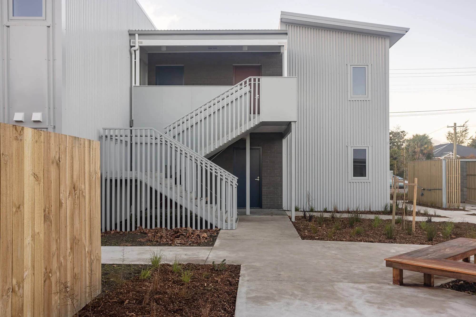 Rear view of a modern multi-story house with white metal siding, an external staircase, and a small patio area with dirt and plants in the foreground.