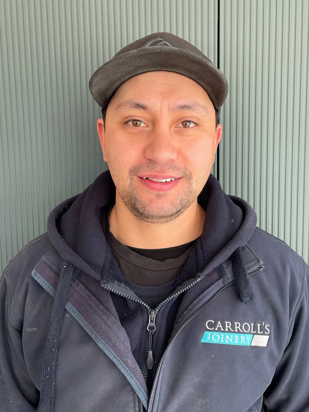 A man wearing a black hoodie and black jacket with a "Carroll's Joinery" logo, and a gray cap, smiling at the camera with a ribbed wall background.