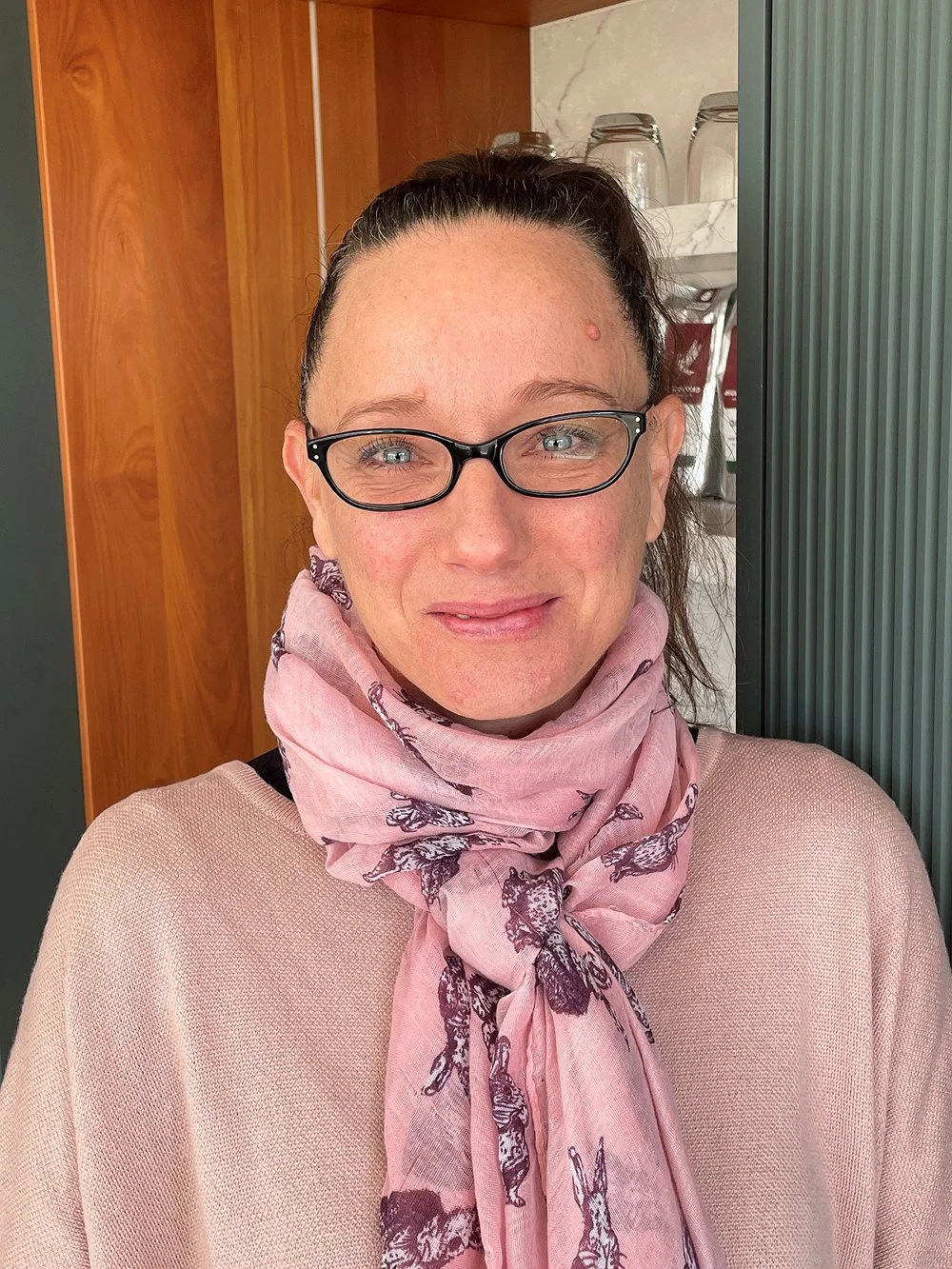 A woman with glasses and a pink butterfly-patterned scarf smiling indoors.