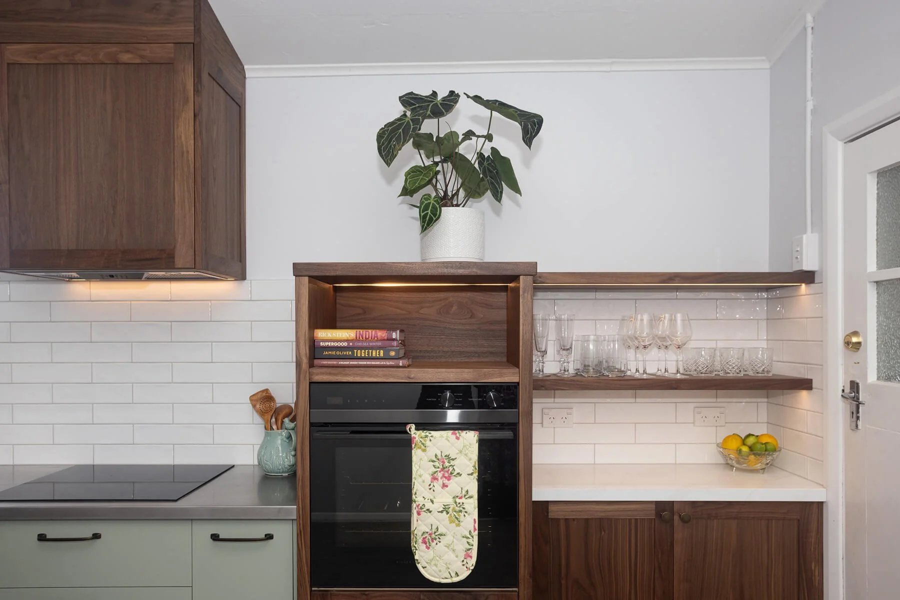 Kitchen with sage green cabinetry, walnut joinery and open glassware shelving.