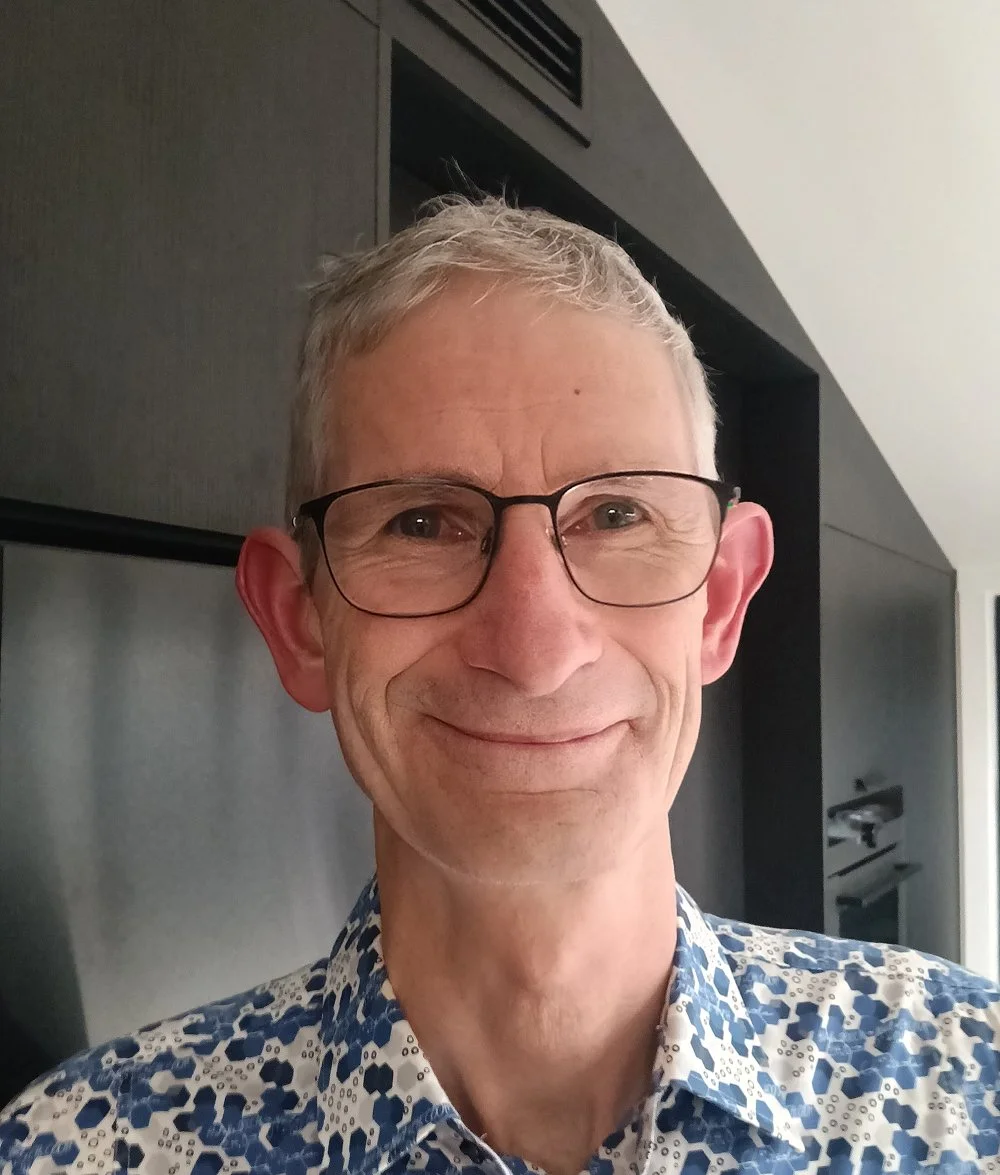 A smiling man with glasses and short gray hair takes a selfie indoors, wearing a patterned blue and white shirt.