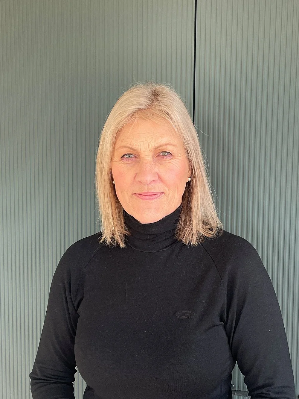 A portrait of a woman with shoulder-length blonde hair, wearing a black turtleneck, standing against a green ribbed metal background.