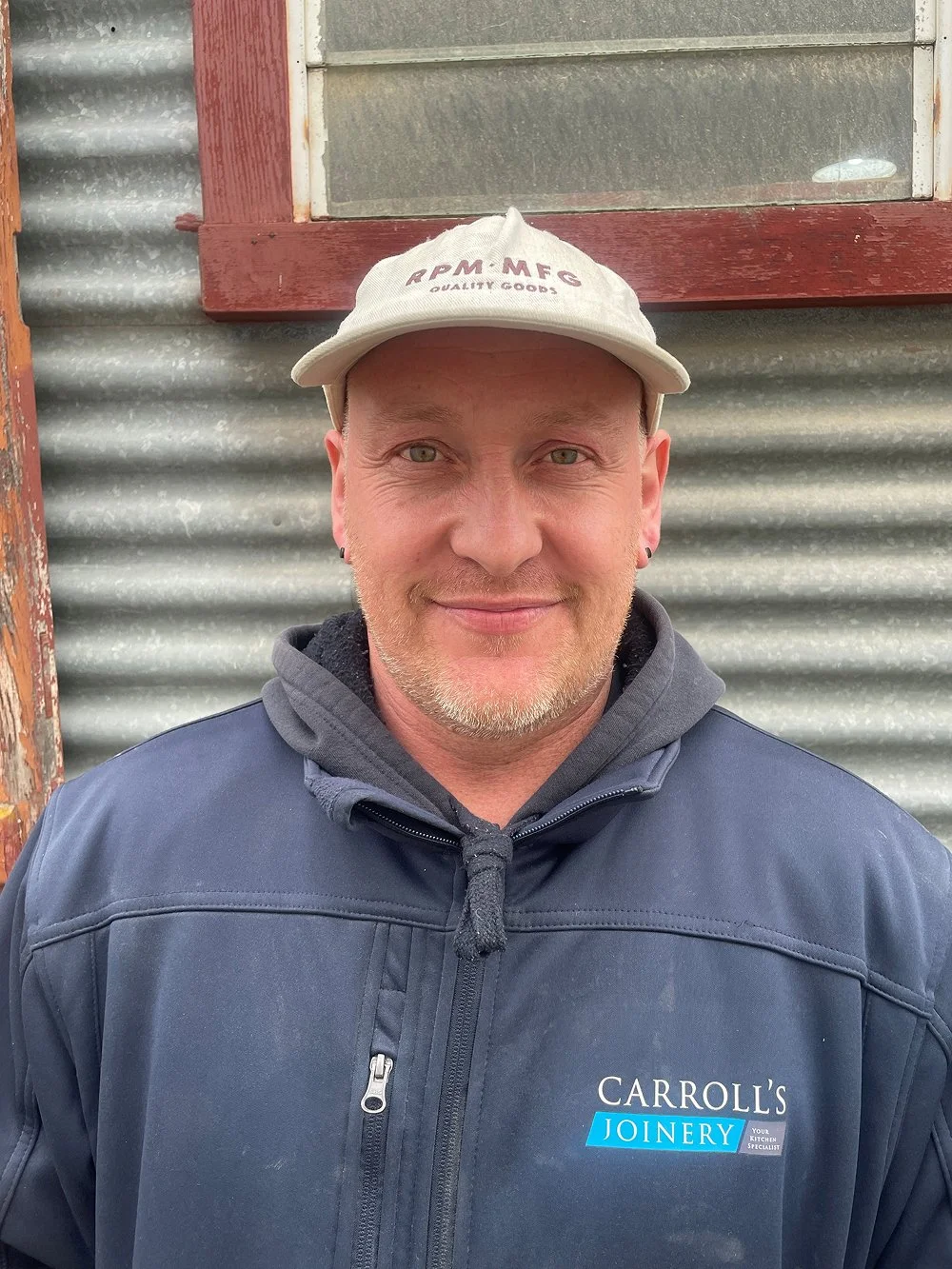Smiling man wearing a gray baseball cap and a navy jacket with a logo that reads Carroll's Joinery, standing in front of a corrugated metal wall with a window.