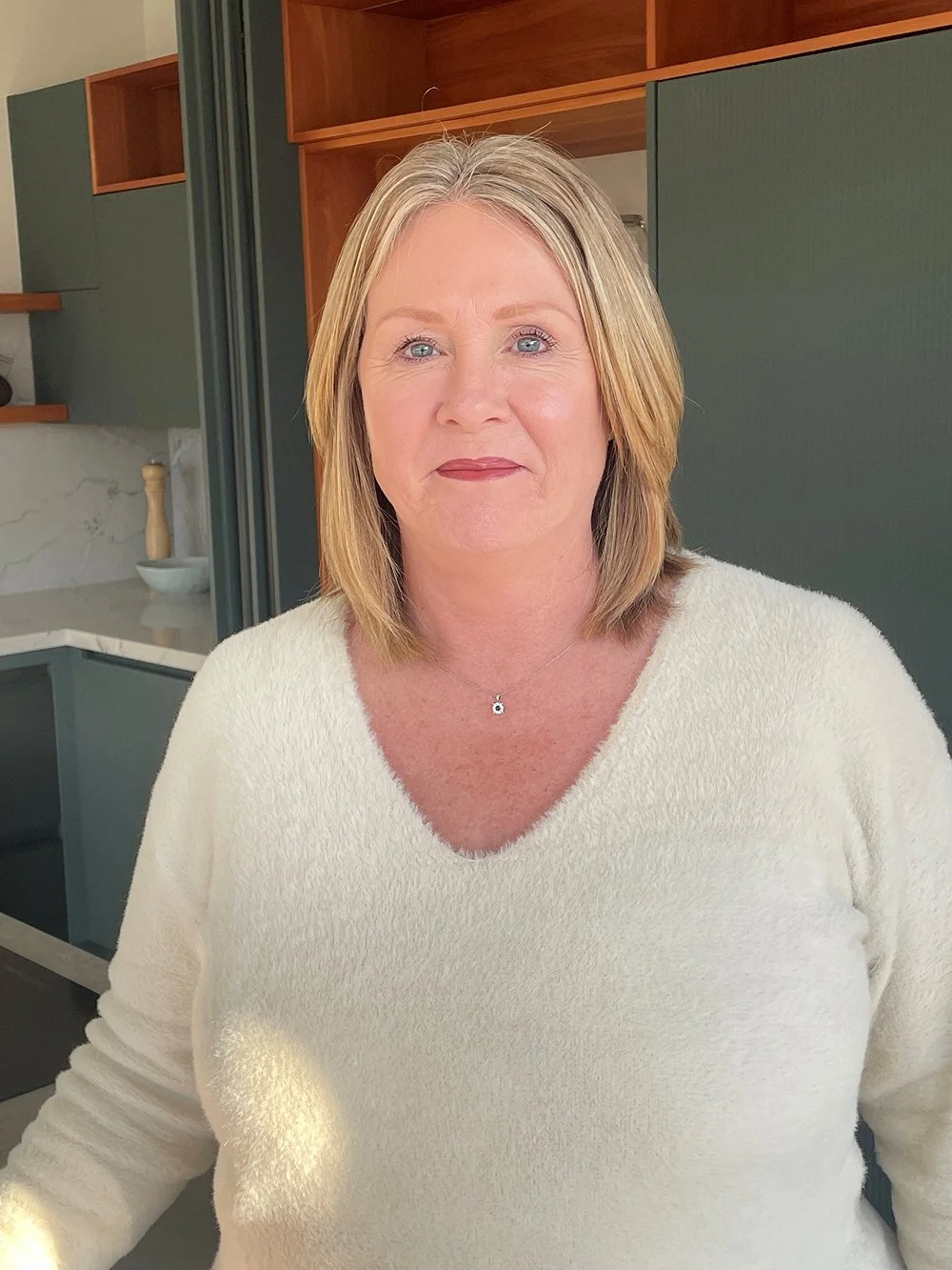 A middle-aged woman with blonde hair and blue eyes, wearing a white sweater and a small necklace, standing in a modern kitchen.