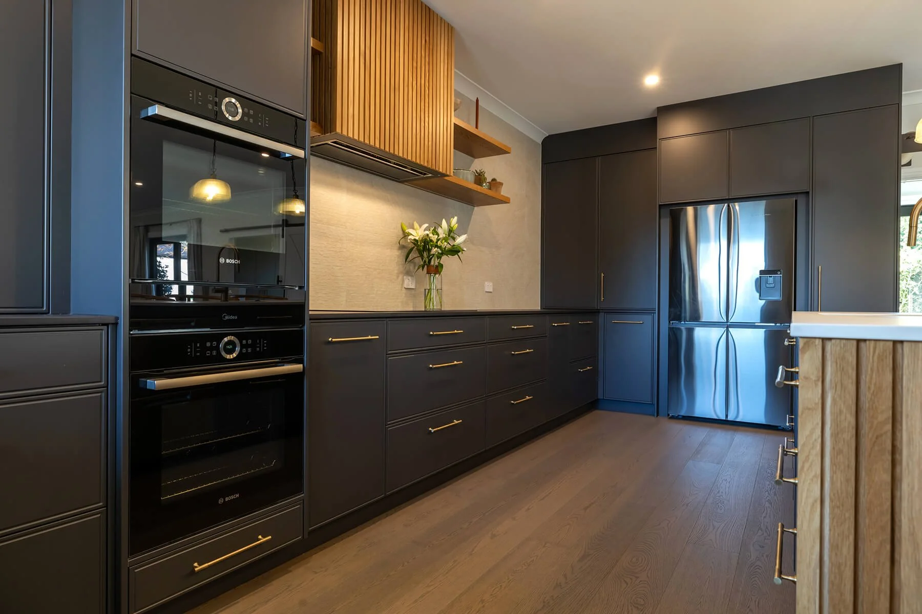 Dark kitchen cabinetry with integrated appliances, timber detailing and brass handles.