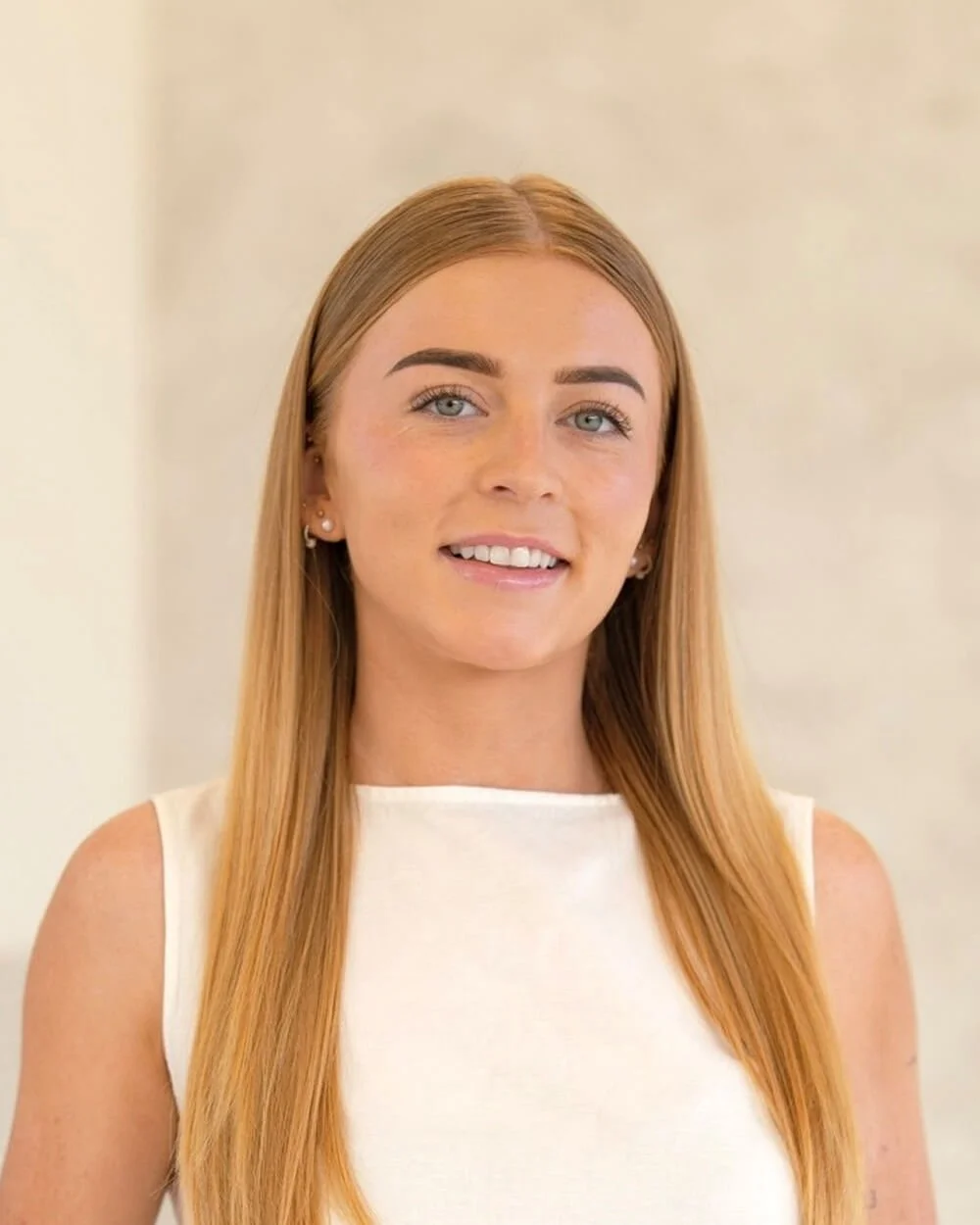 A young woman with long, straight, blonde hair, wearing a sleeveless white top and light blue jeans, standing indoors against a neutral background.