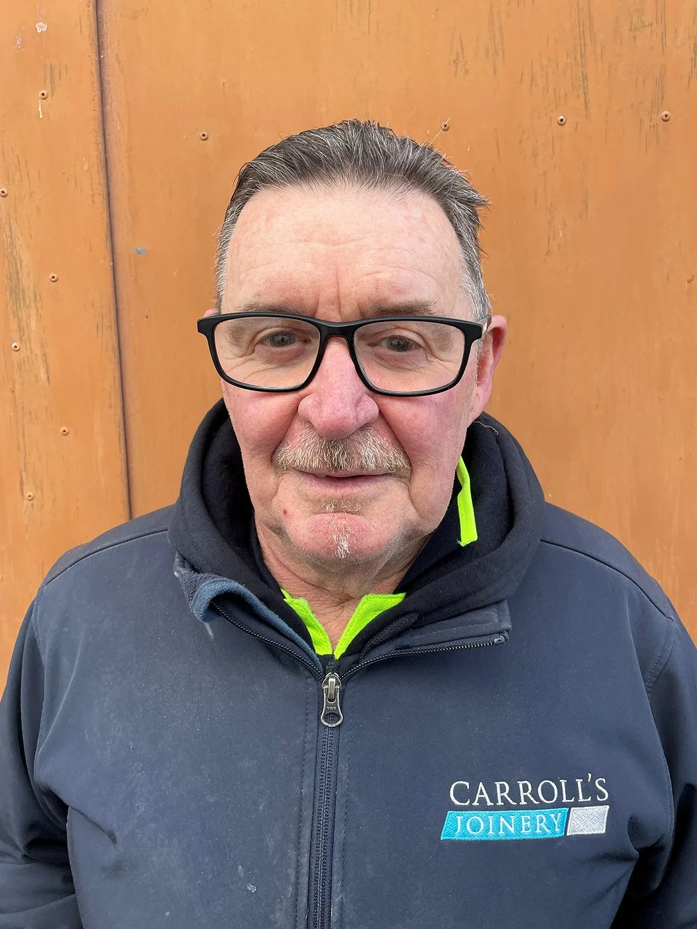 A middle-aged man with glasses, short dark hair, and a mustache standing in front of a wooden background, wearing a navy blue jacket with the logo 'Carroll's Joinery'.