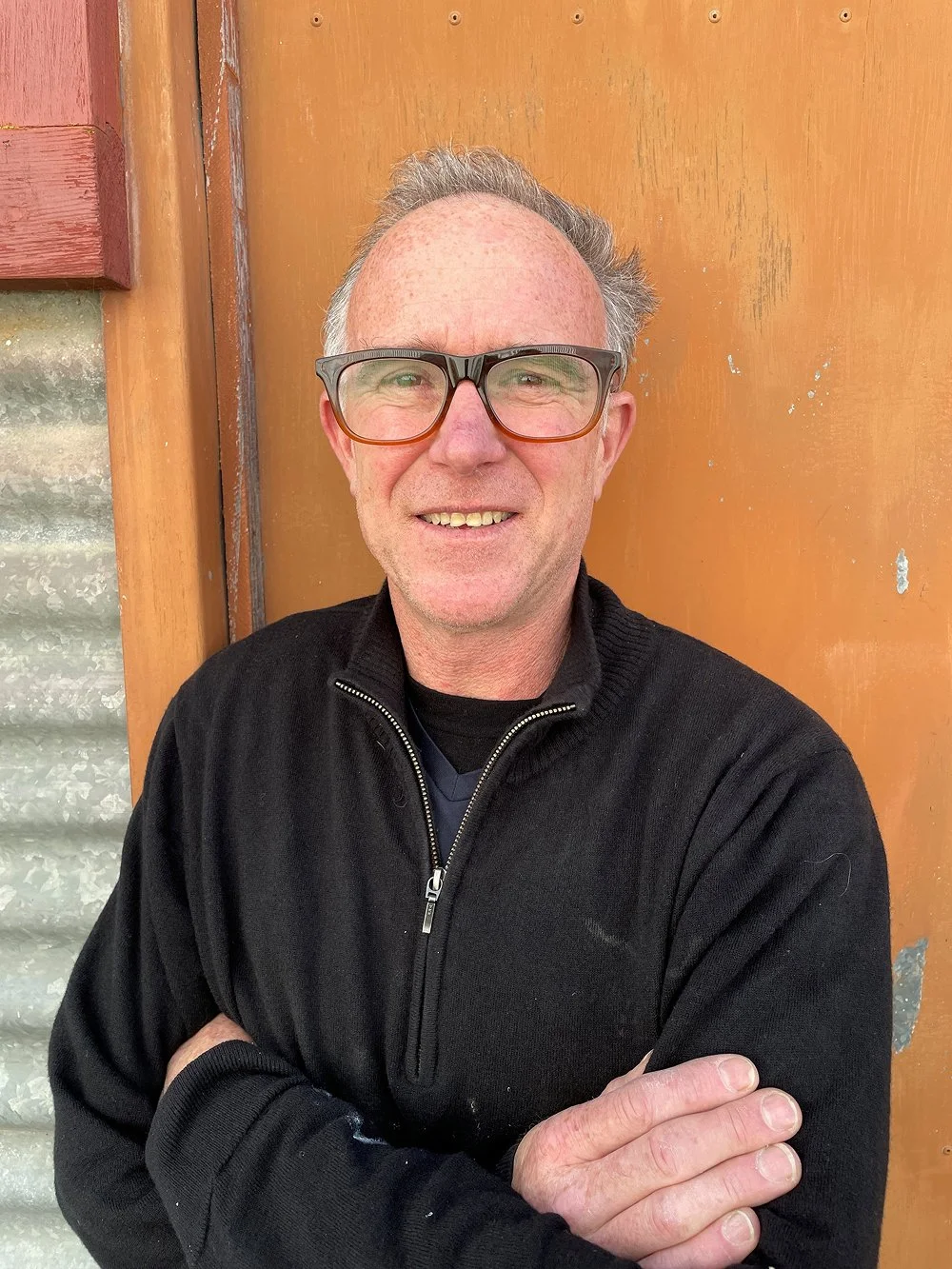 A middle-aged man with gray hair, wearing glasses and a black zip-up sweater, standing against a wooden background with metal siding visible on the left.