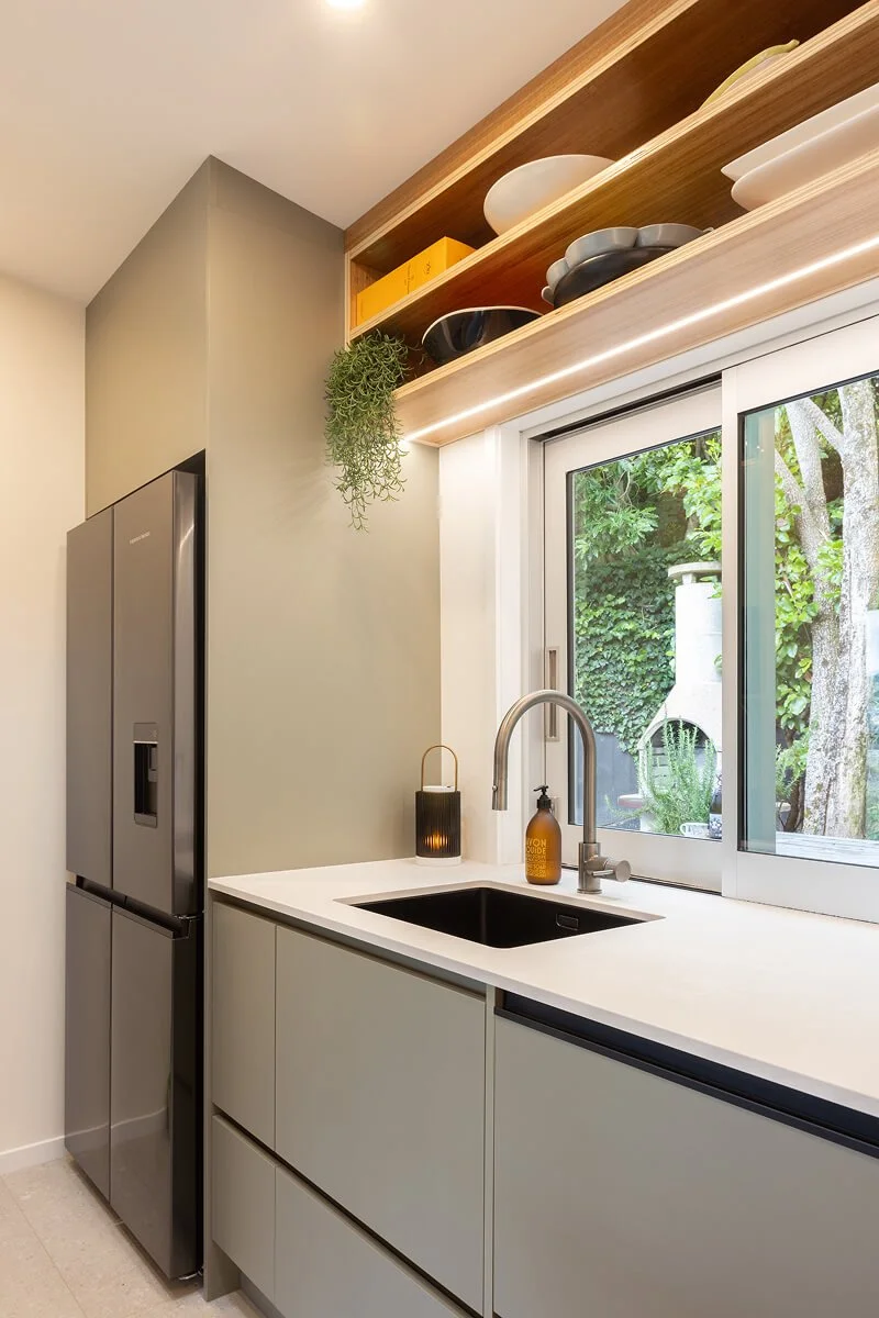 Compact kitchen with sage cabinetry, timber shelving and open storage above the sink.