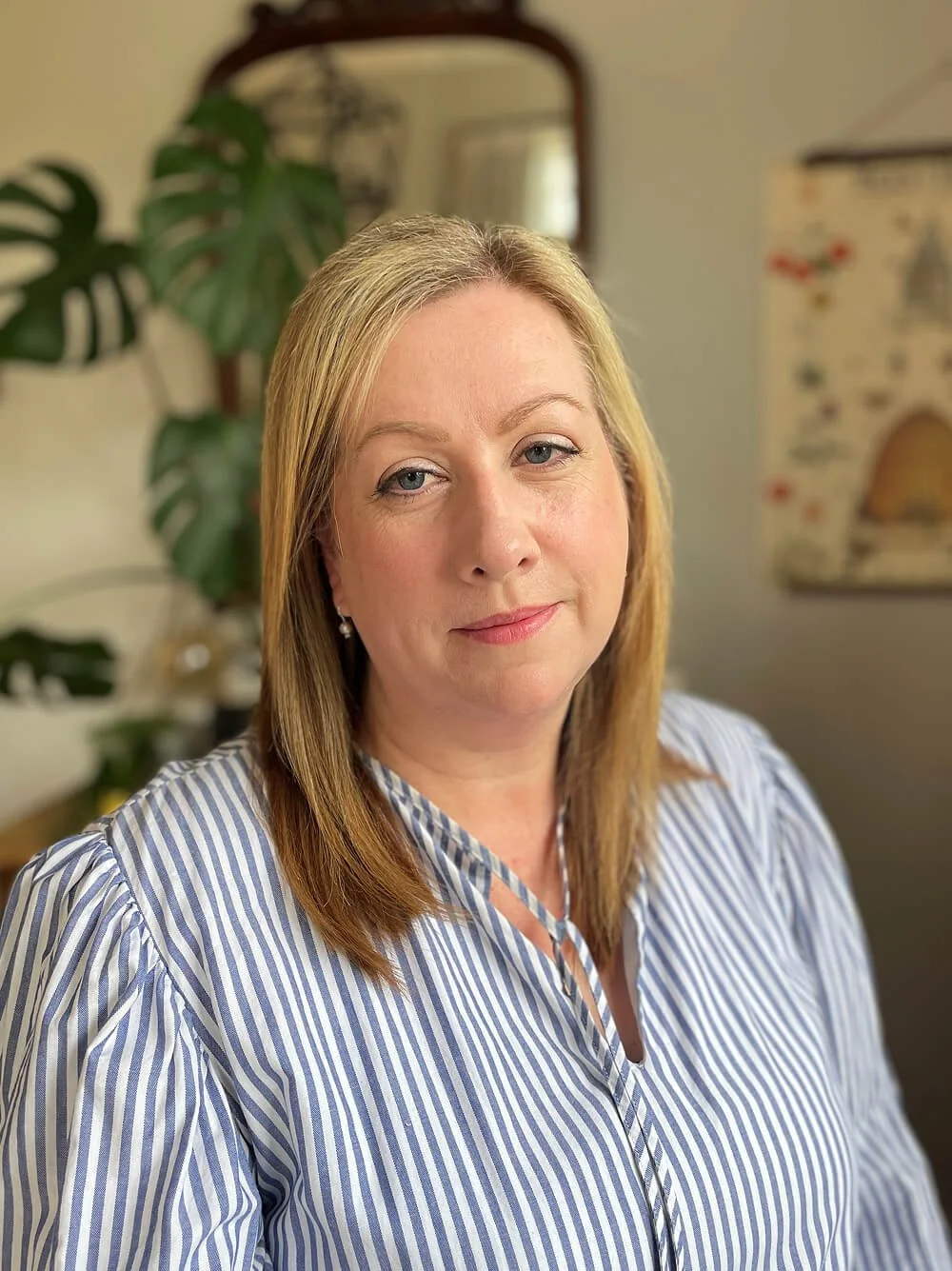 Portrait of a woman with blonde hair wearing a blue and white striped blouse, indoors with a plant and mirror in the background.