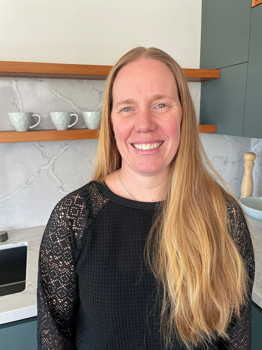 A woman with long red hair and a black lace top smiling in a modern kitchen with white marble backsplash and three white coffee cups on a wooden shelf.